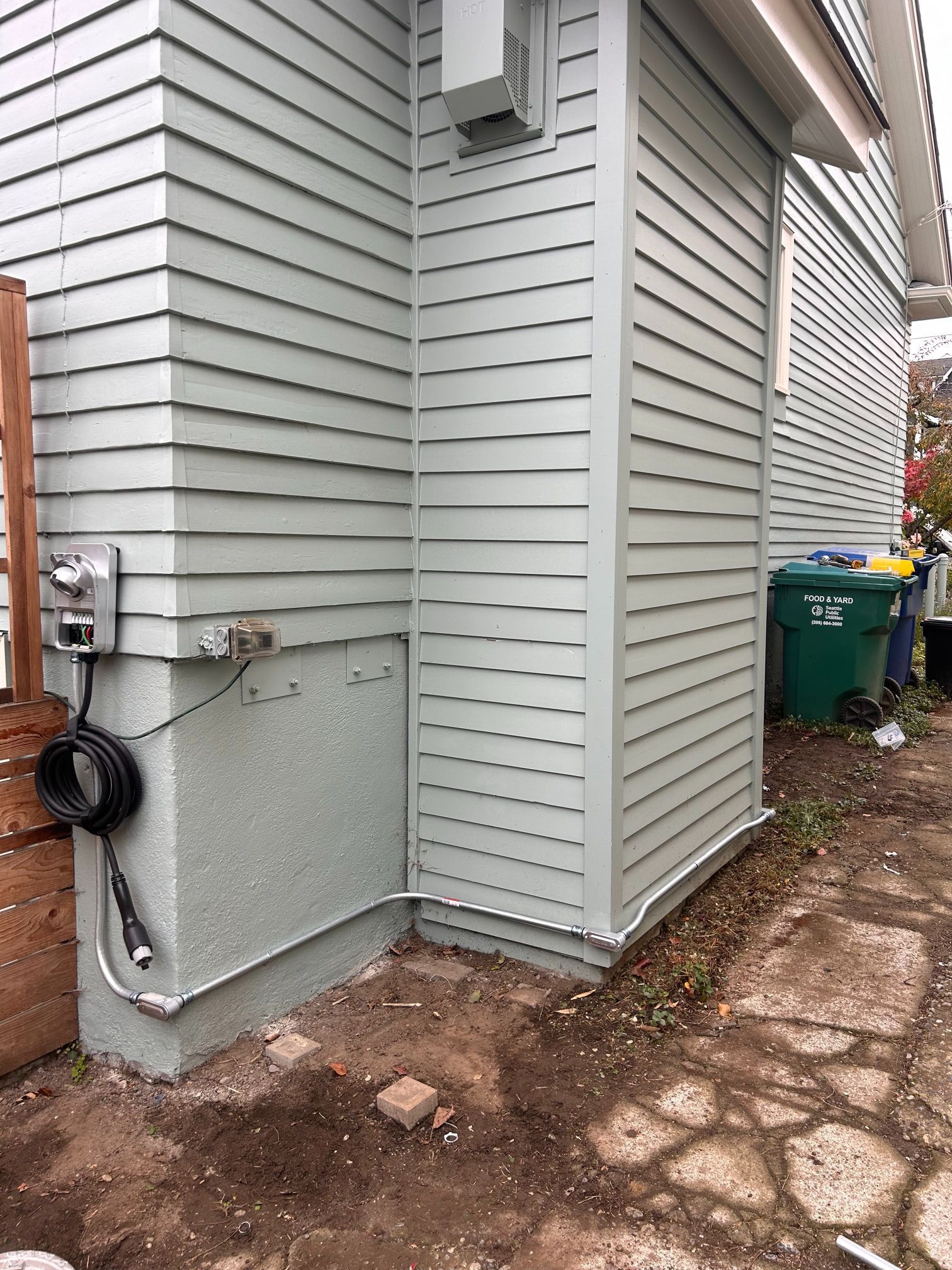 Side of a gray house with utility meter, hoses, and a dirt path beside a narrow yard