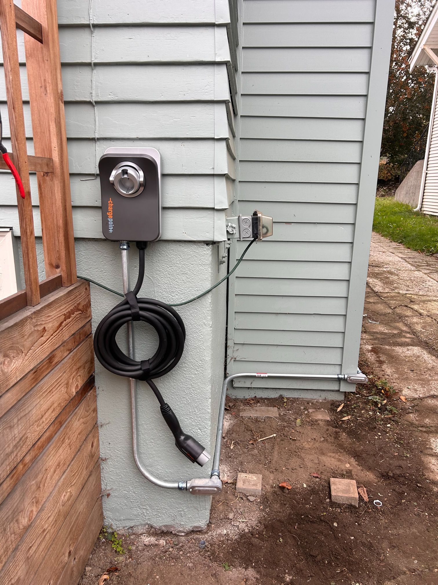Electric meter and conduit mounted on a house exterior with coiled black cable near the foundation.