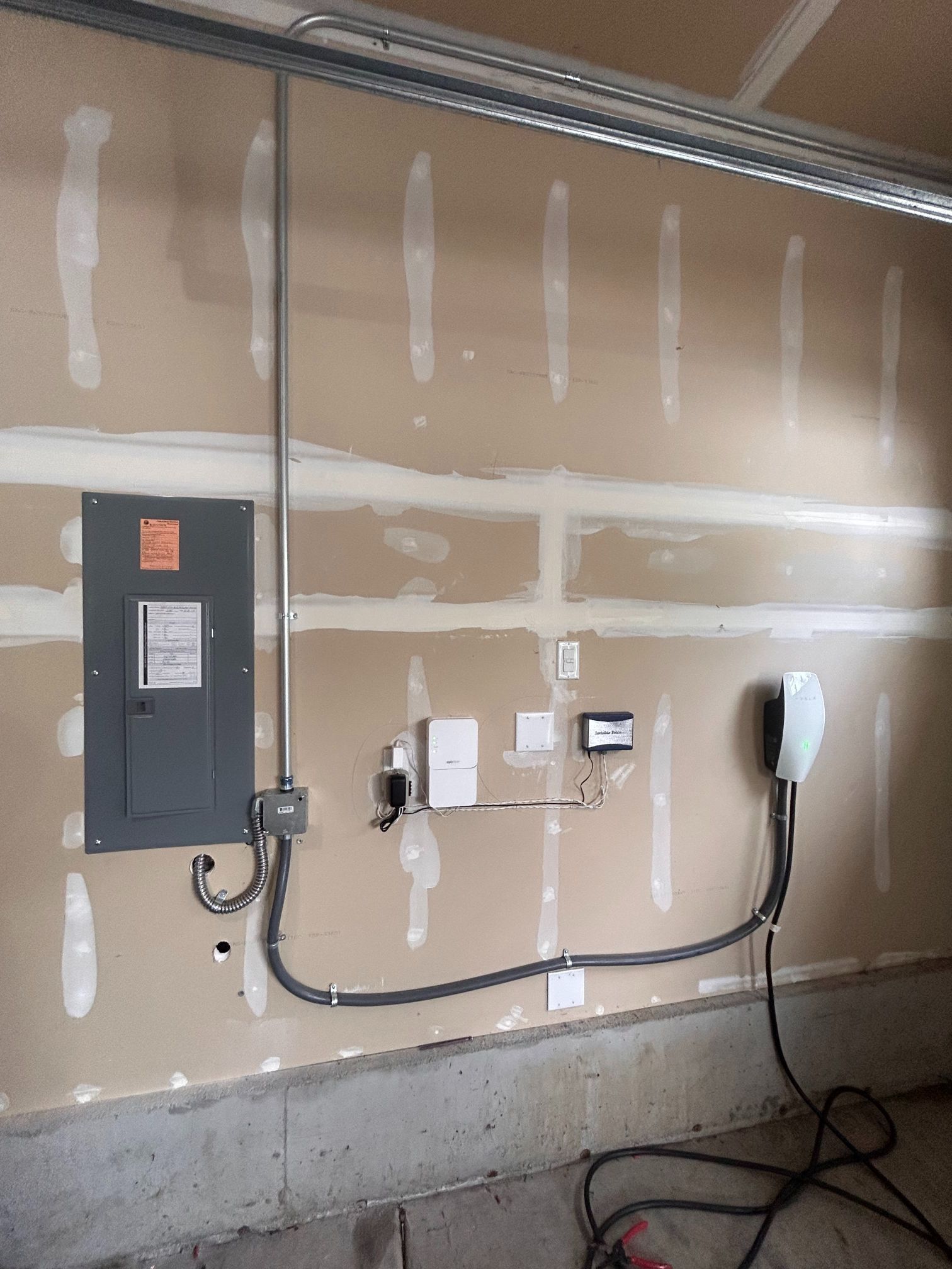 Garage wall with mounted electrical panel, outlet, EV charger, and exposed cables on unfinished drywall.