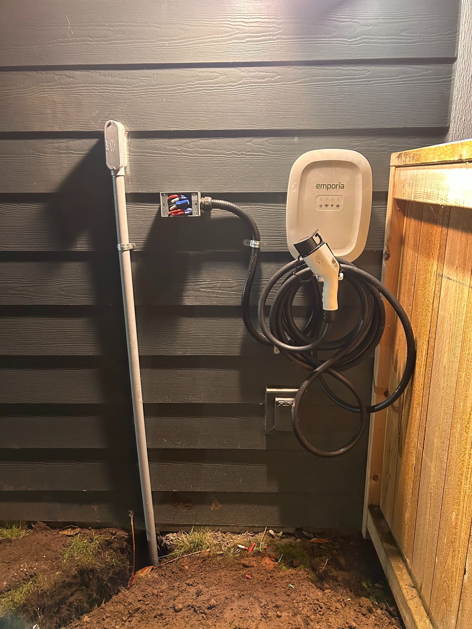 Outdoor wall-mounted electrical outlet with a plugged-in cable beside a wooden structure.