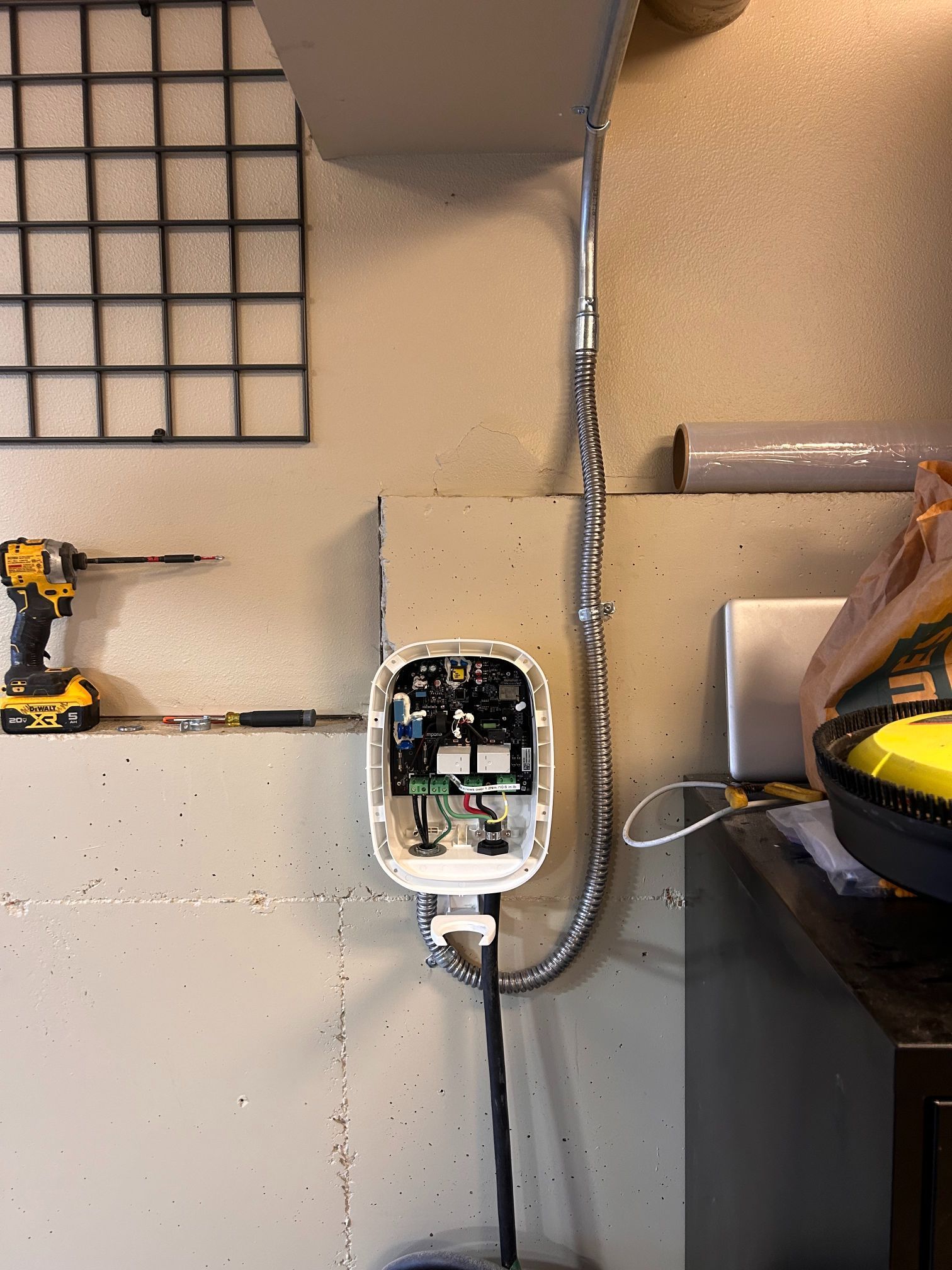 Open electrical junction box with exposed wiring on a workshop wall beside a drill and tool bench.