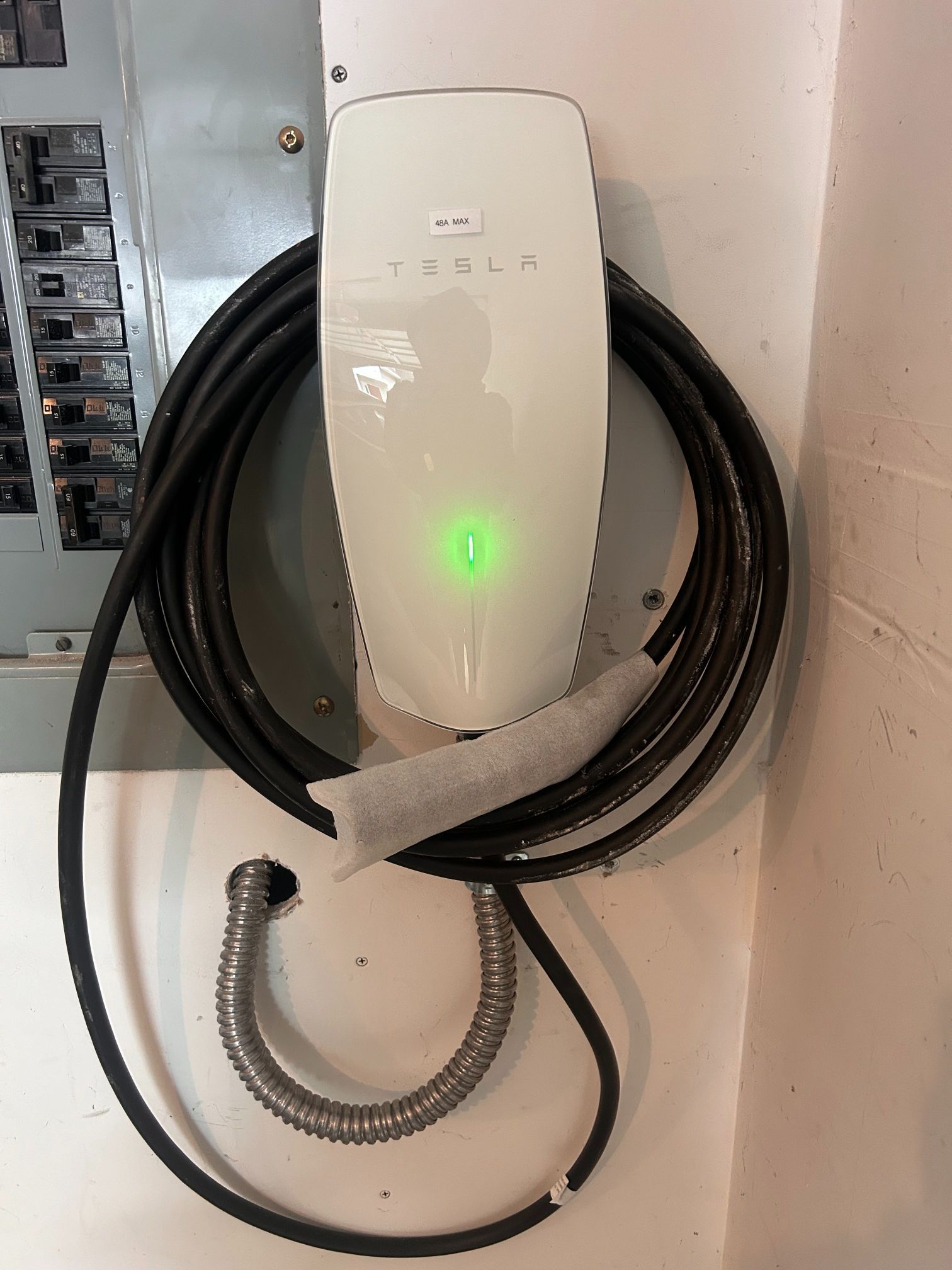 Wall-mounted Tesla charging unit with a glowing green indicator and coiled cables beside an electrical panel.
