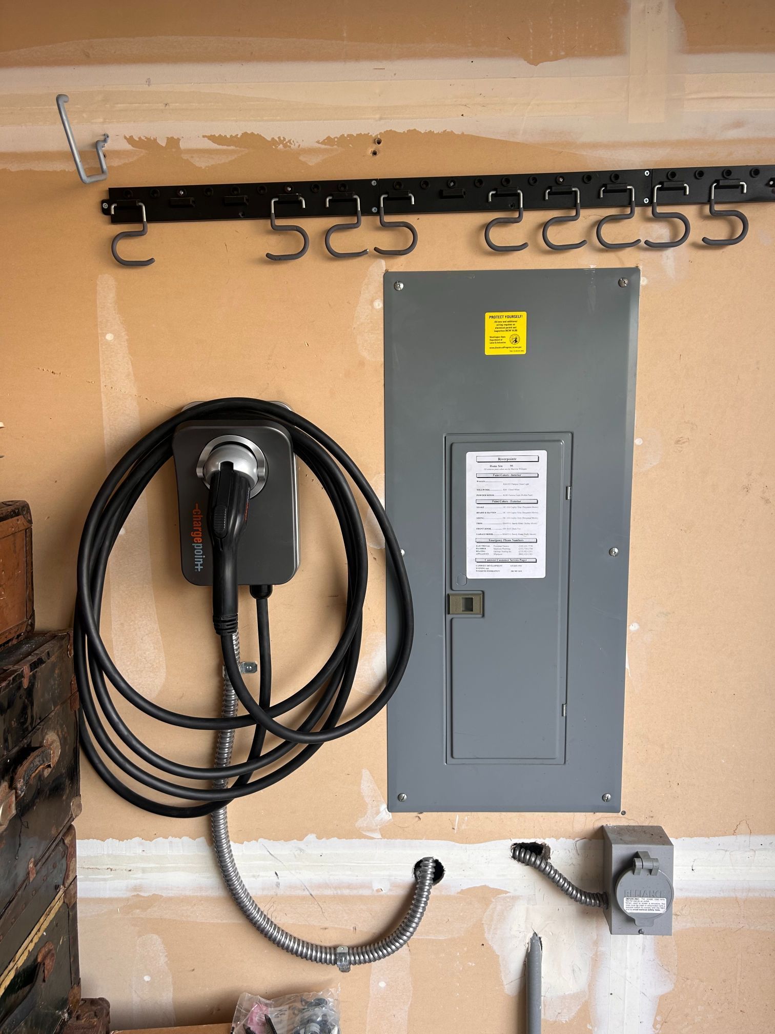 Wall-mounted electrical panel with coiled charging cable, hooks, and outlet on a garage wall.