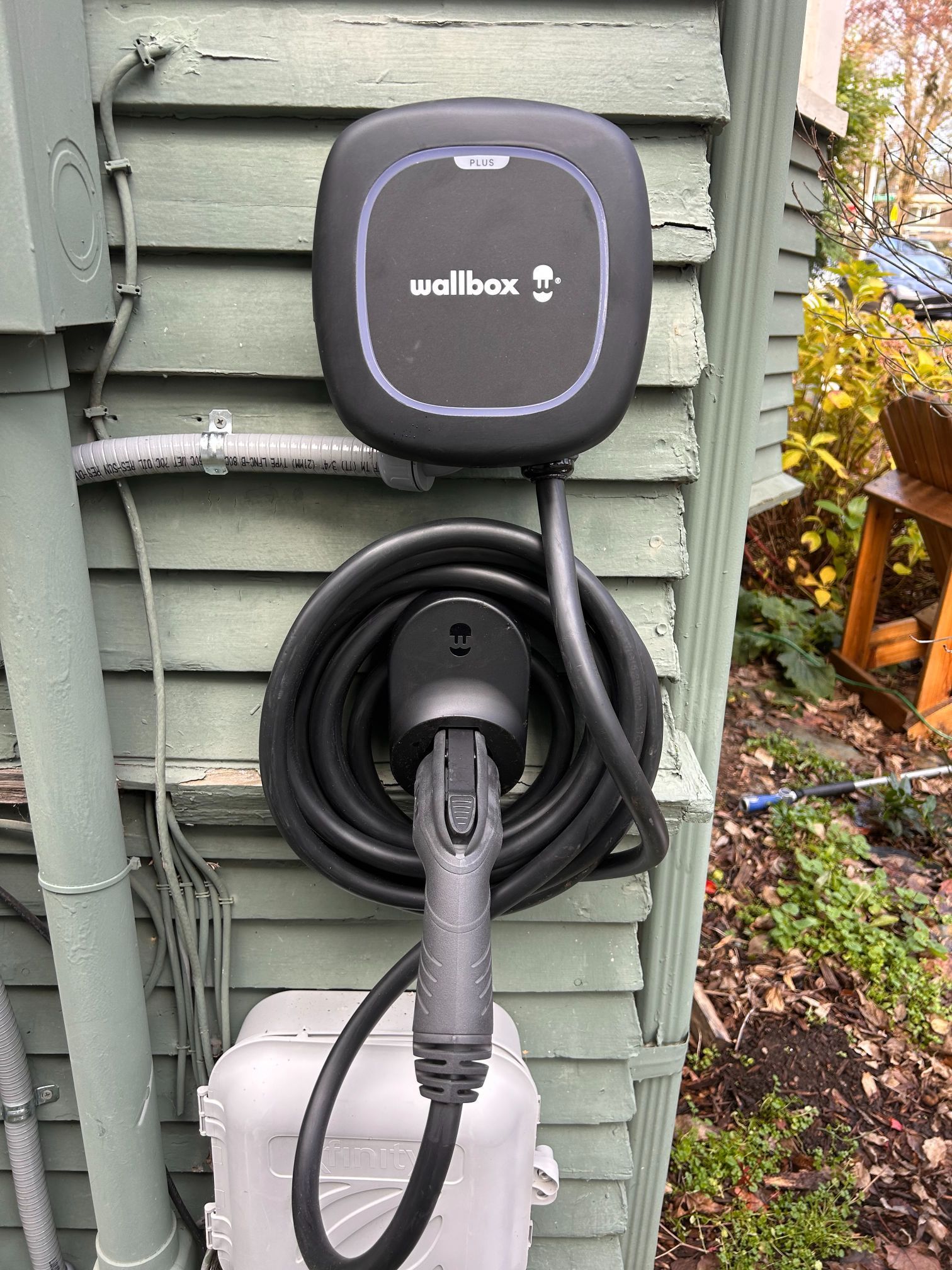 Wallbox EV charger mounted on green siding with coiled black charging cable and plug.