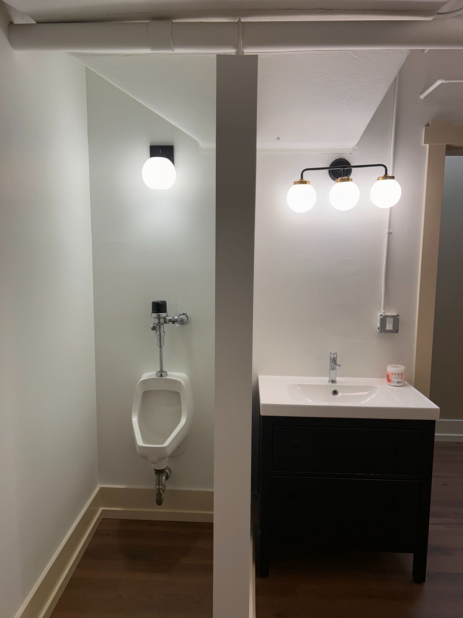Public restroom with urinal, toilet stall partition, and sink with lights above.