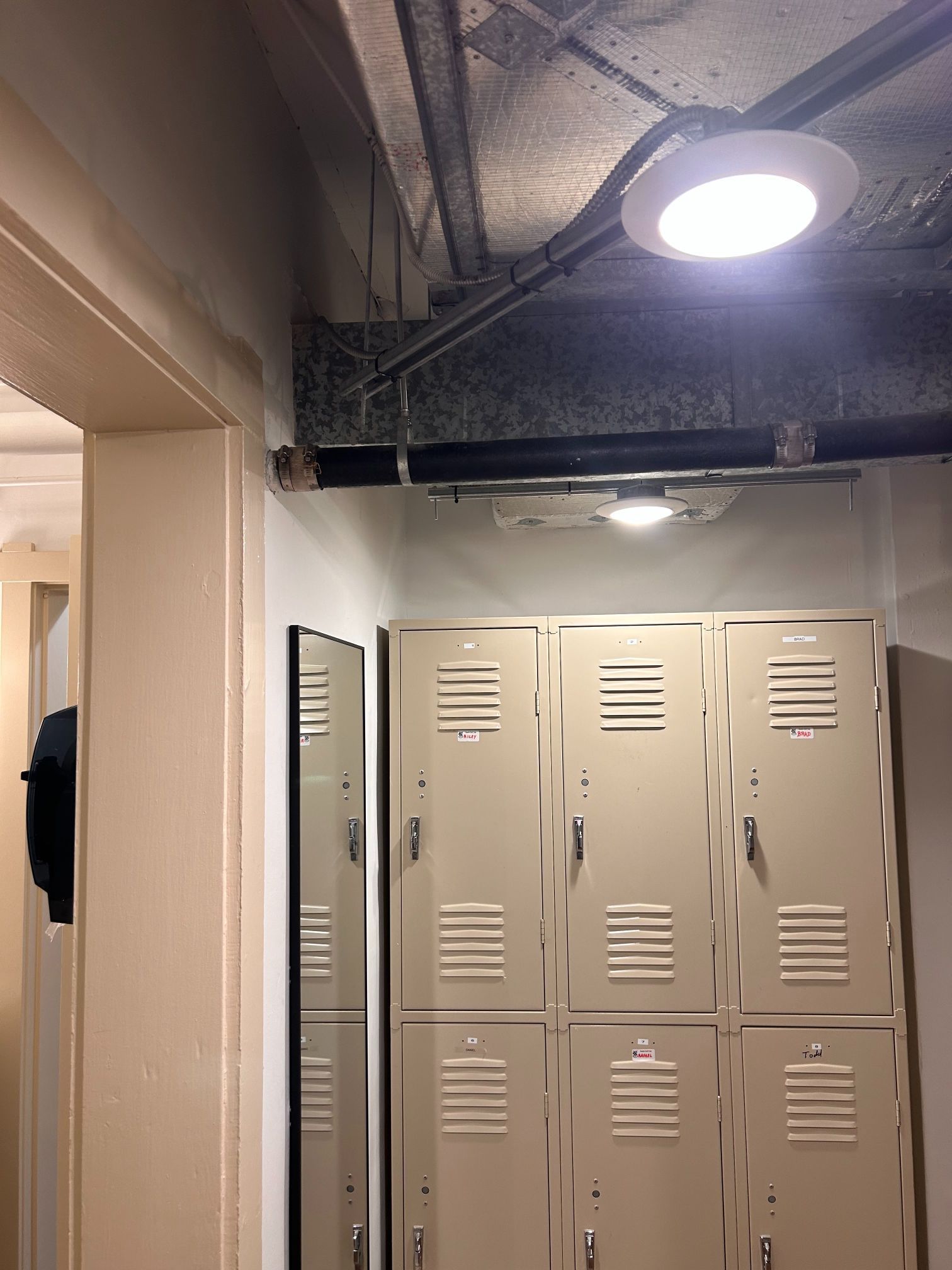 Dim hallway with beige lockers and exposed ceiling pipes under a bright round light.