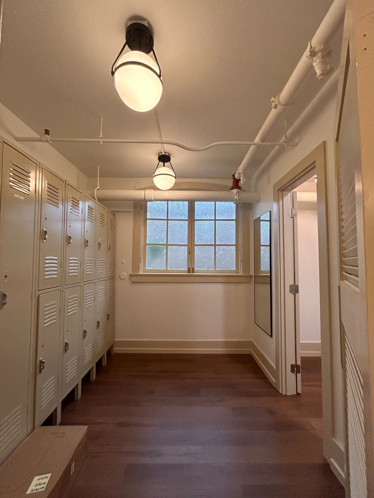 Narrow locker-lined hallway with white walls, ceiling lights, and a window at the far end.