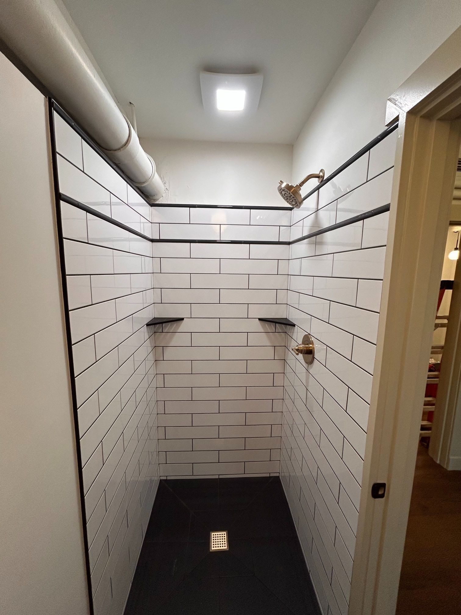 Small tiled shower stall with white brick-style walls, black floor, and wall shelves under a ceiling light