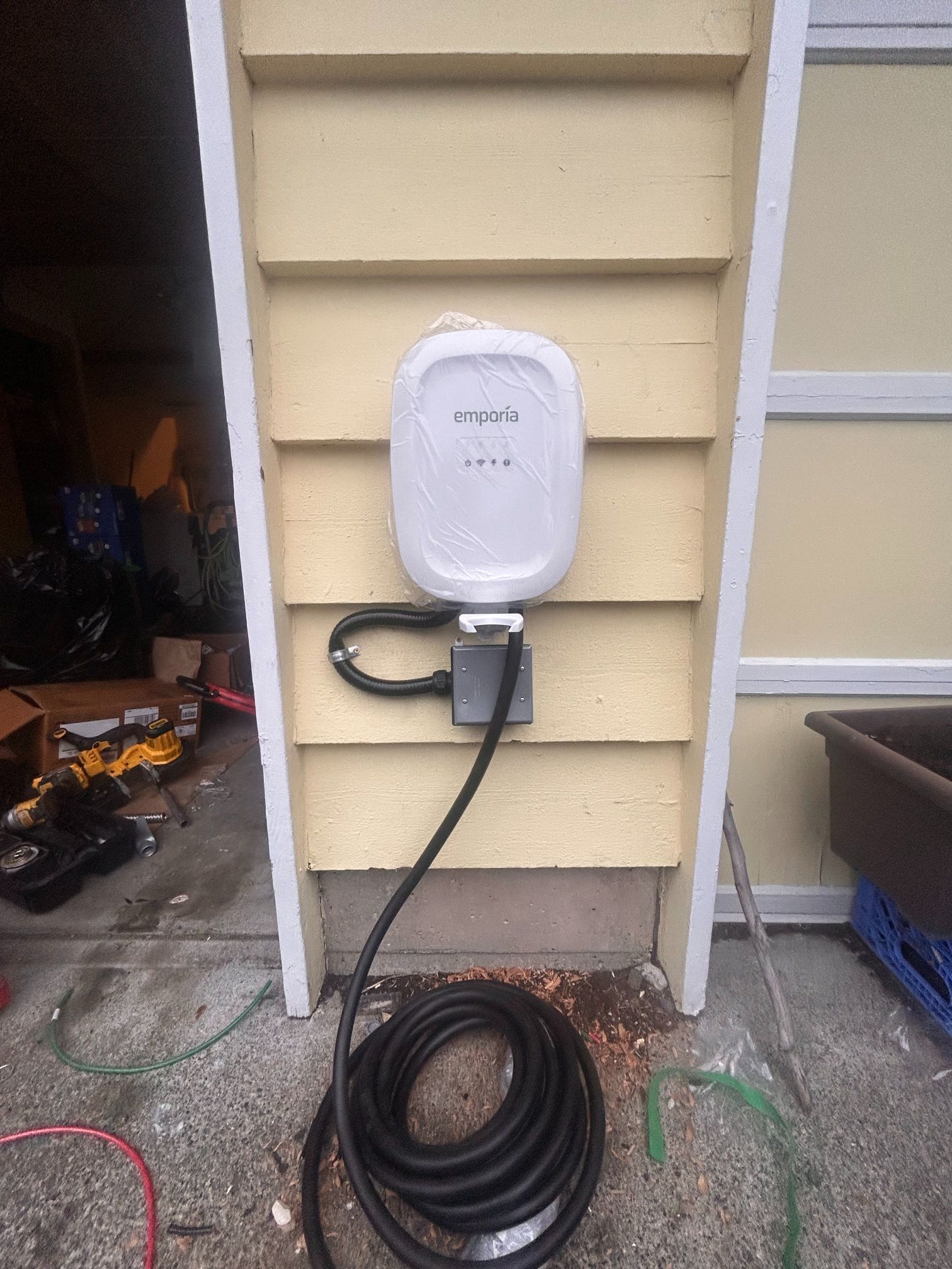EV charger mounted on beige house siding with coiled cable on the ground near a garage.