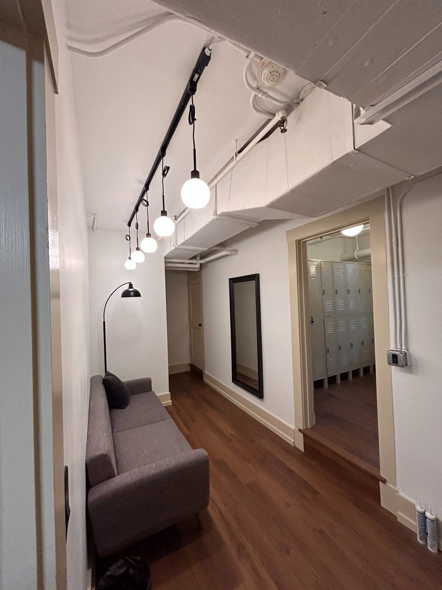 Long hallway with wood floors, gray sofa, hanging lights, and mirrored office doors
