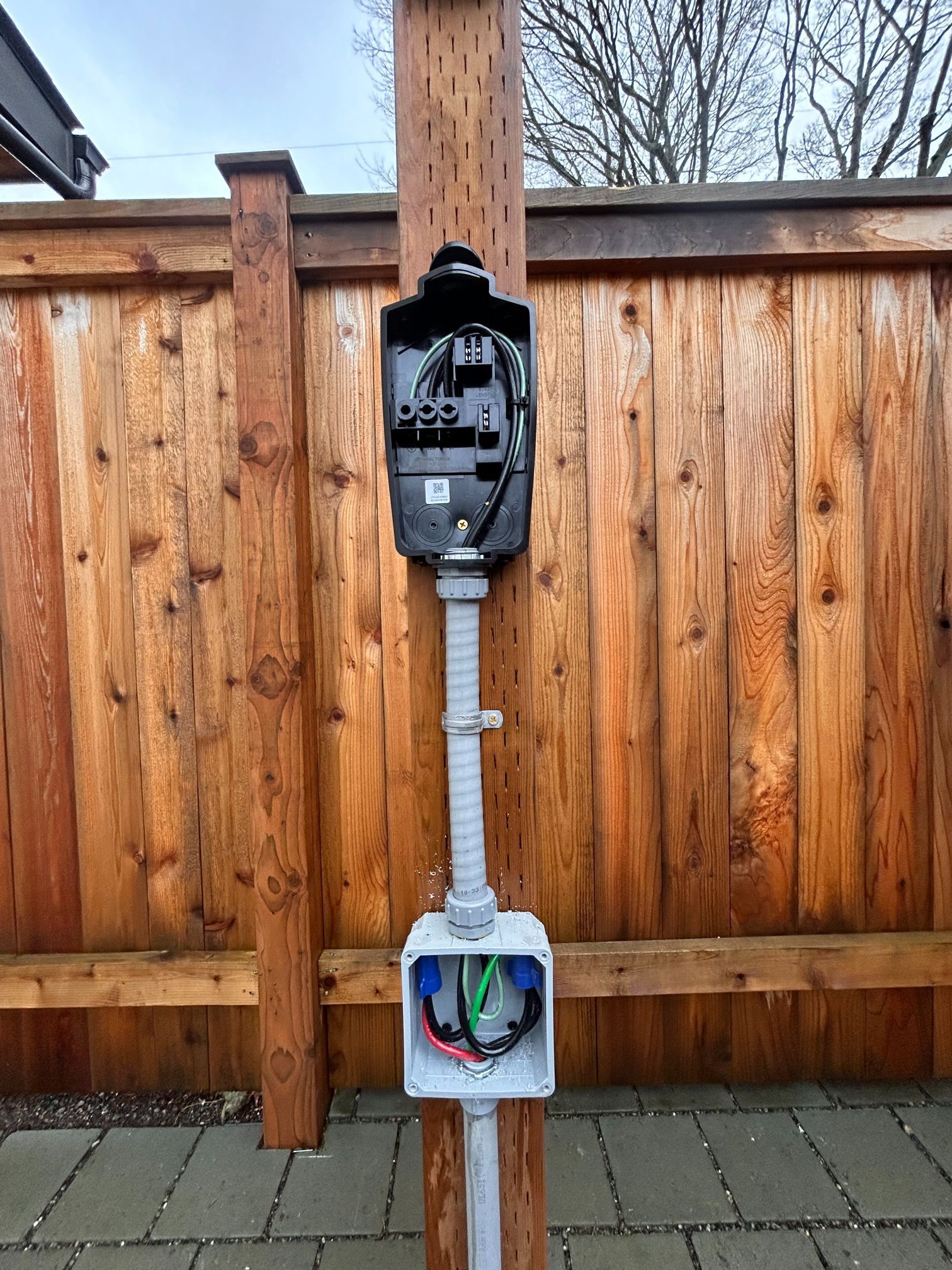 Outdoor electrical meter and open utility box mounted on a wooden fence, with exposed wires below.