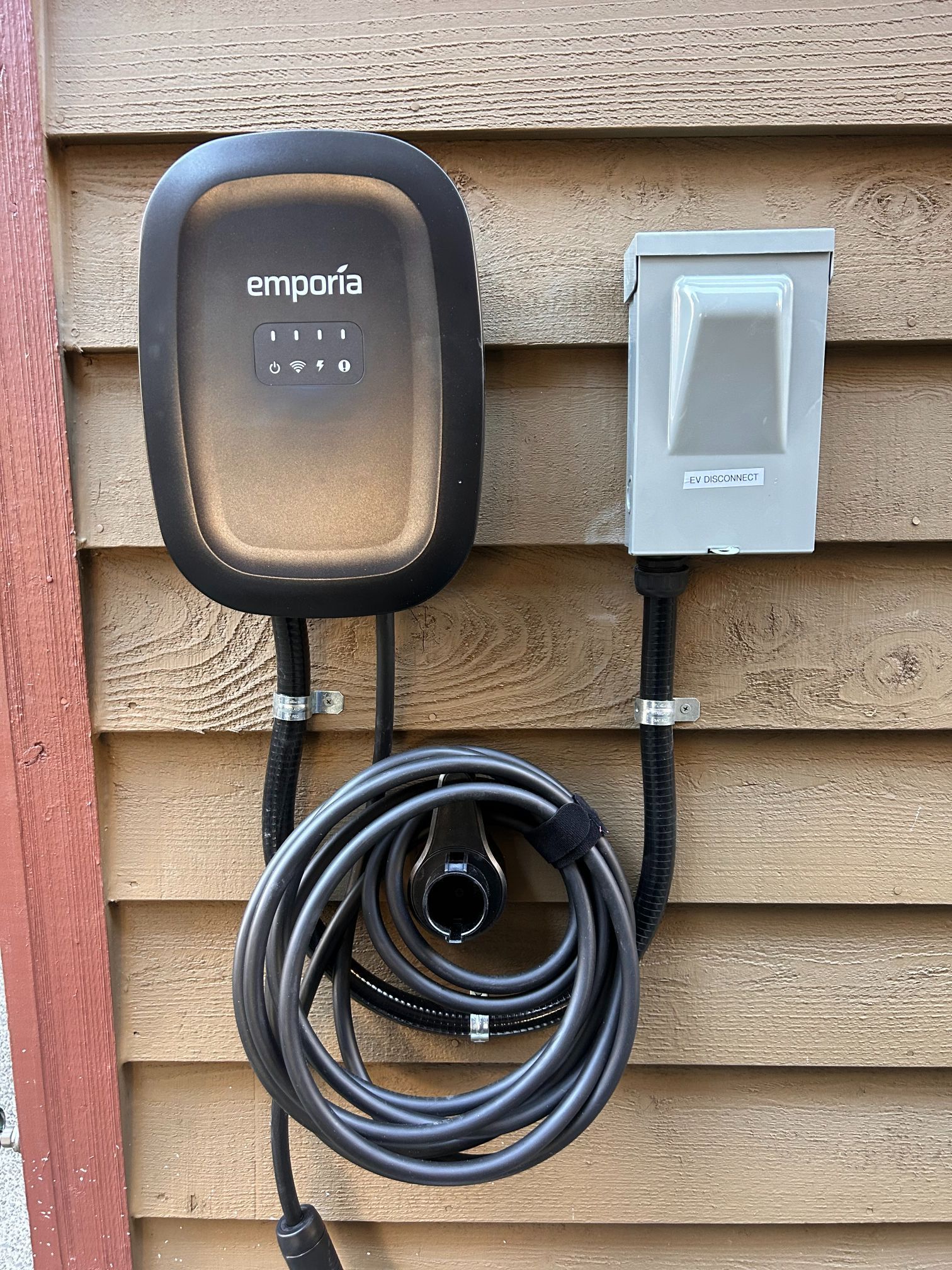 Outdoor Emporia EV charger mounted on a brick wall beside an electrical disconnect box with coiled cable.
