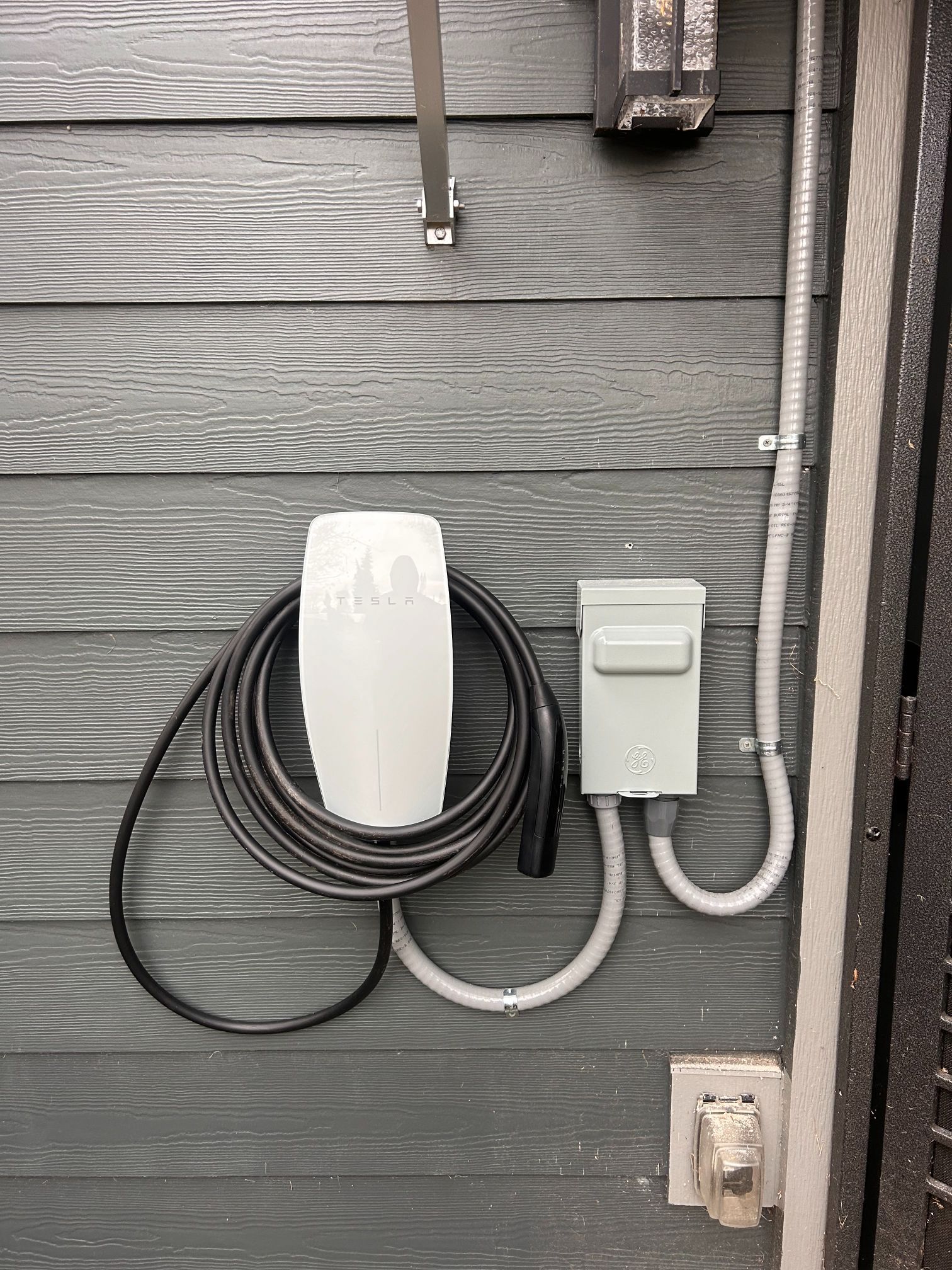 Wall-mounted white EV charger with coiled black cable on gray siding near an outdoor outlet.