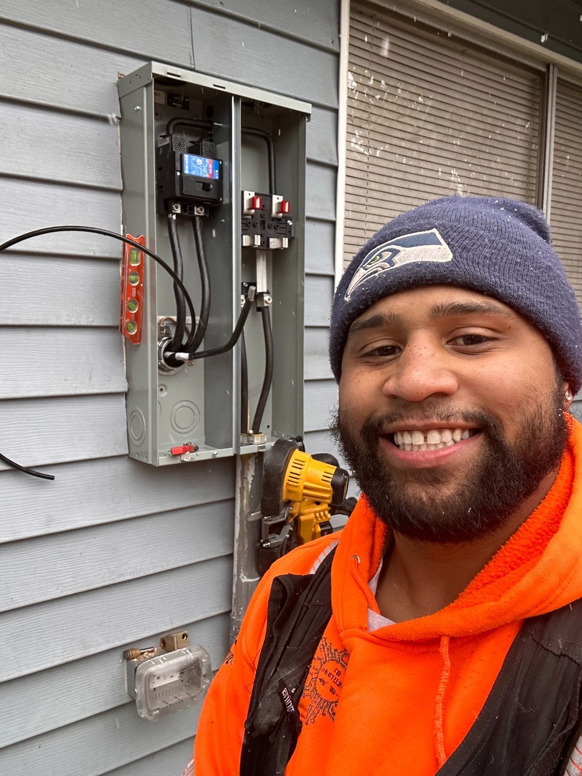 Smiling person in orange hoodie and beanie beside an open electrical panel on a house exterior