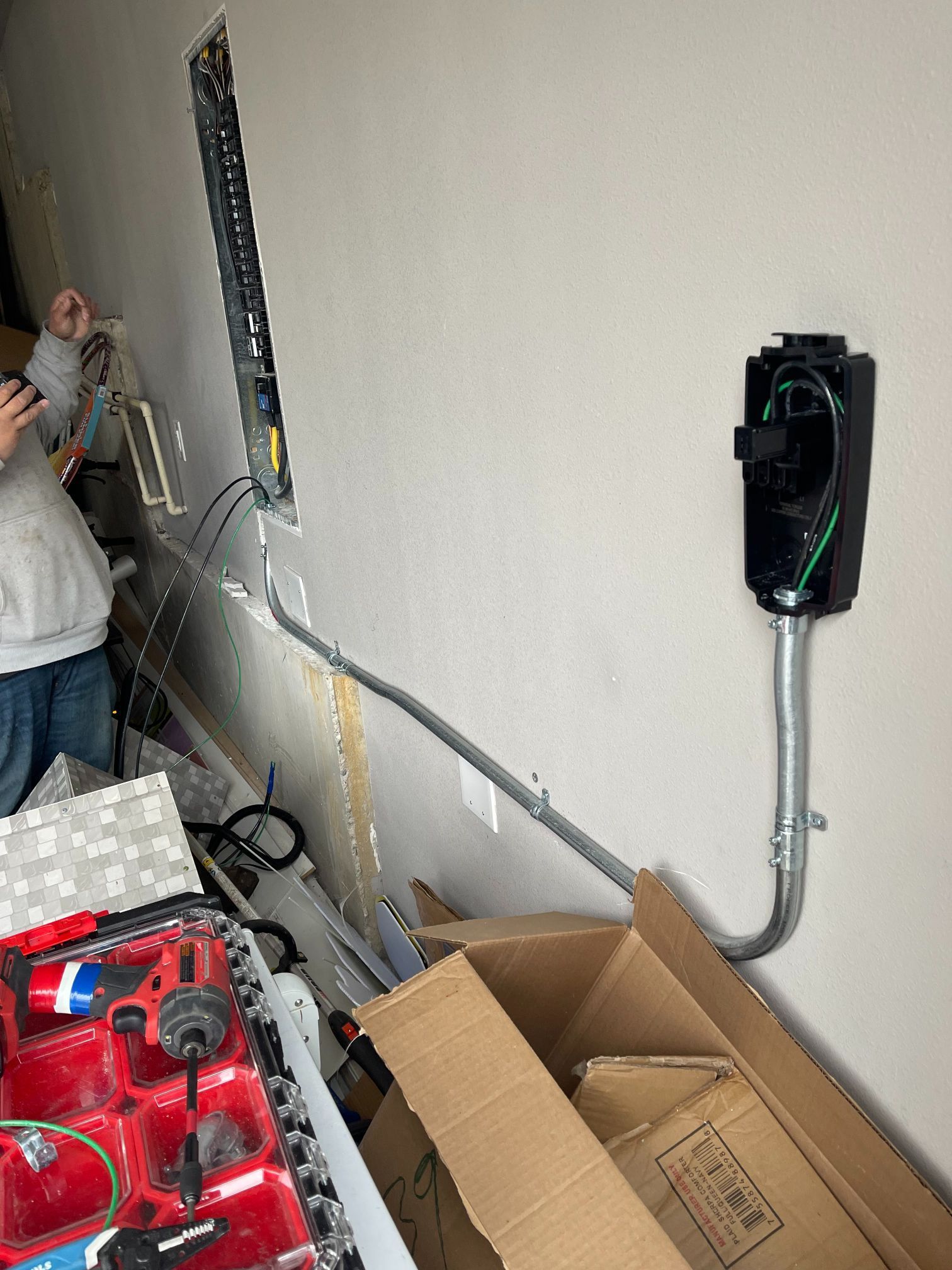Person working in a cluttered room beside a wall-mounted device and exposed wiring.