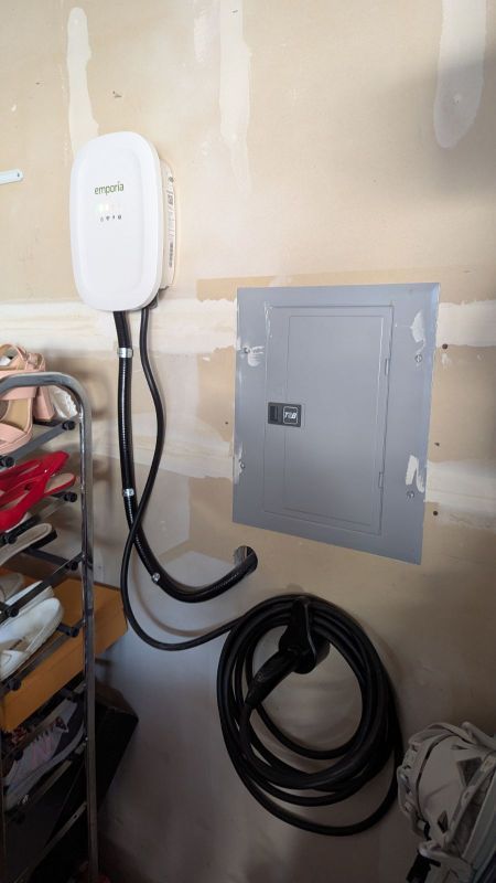 Wall-mounted white EV charger beside a gray electrical panel, with coiled black cable in a garage/workshop.