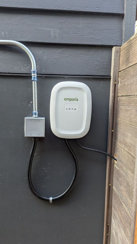 White wall-mounted device with black cable and gray conduit on a dark exterior wall beside a fence