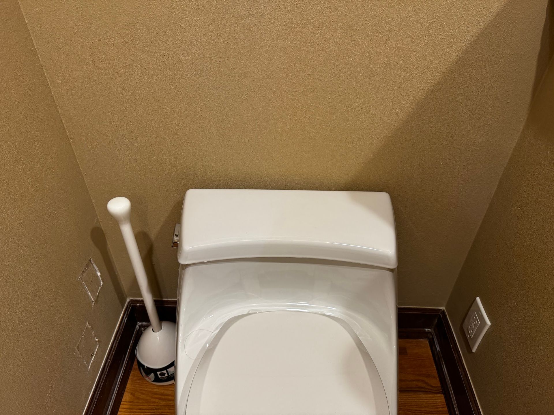 Toilet in a small bathroom with light brown walls and a plunger.