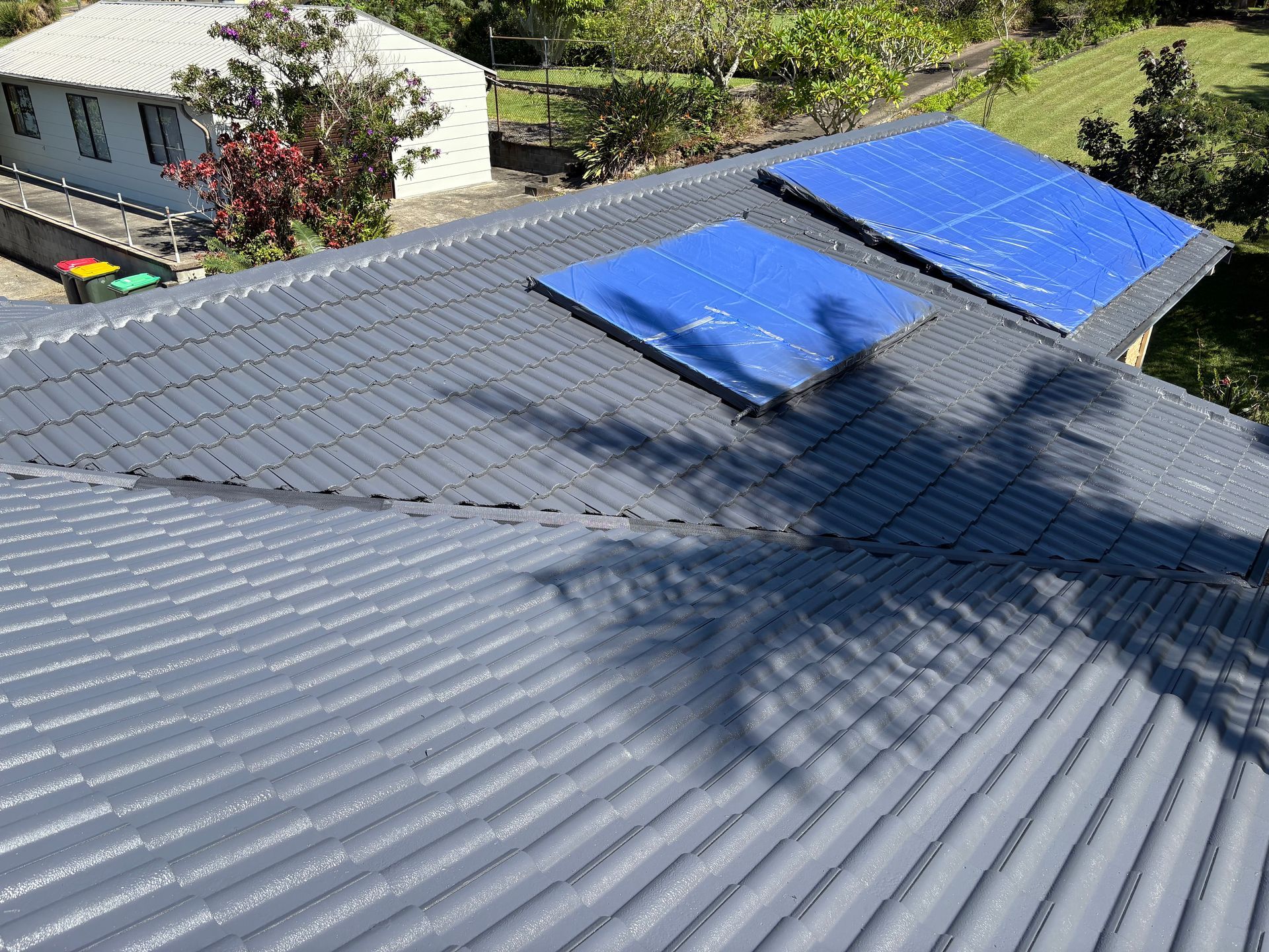 Solar panels on a dark metal roof with a skylight, viewed from above, with a tree shadow across the surface — In-Time Roofing In Newcastle, NSW