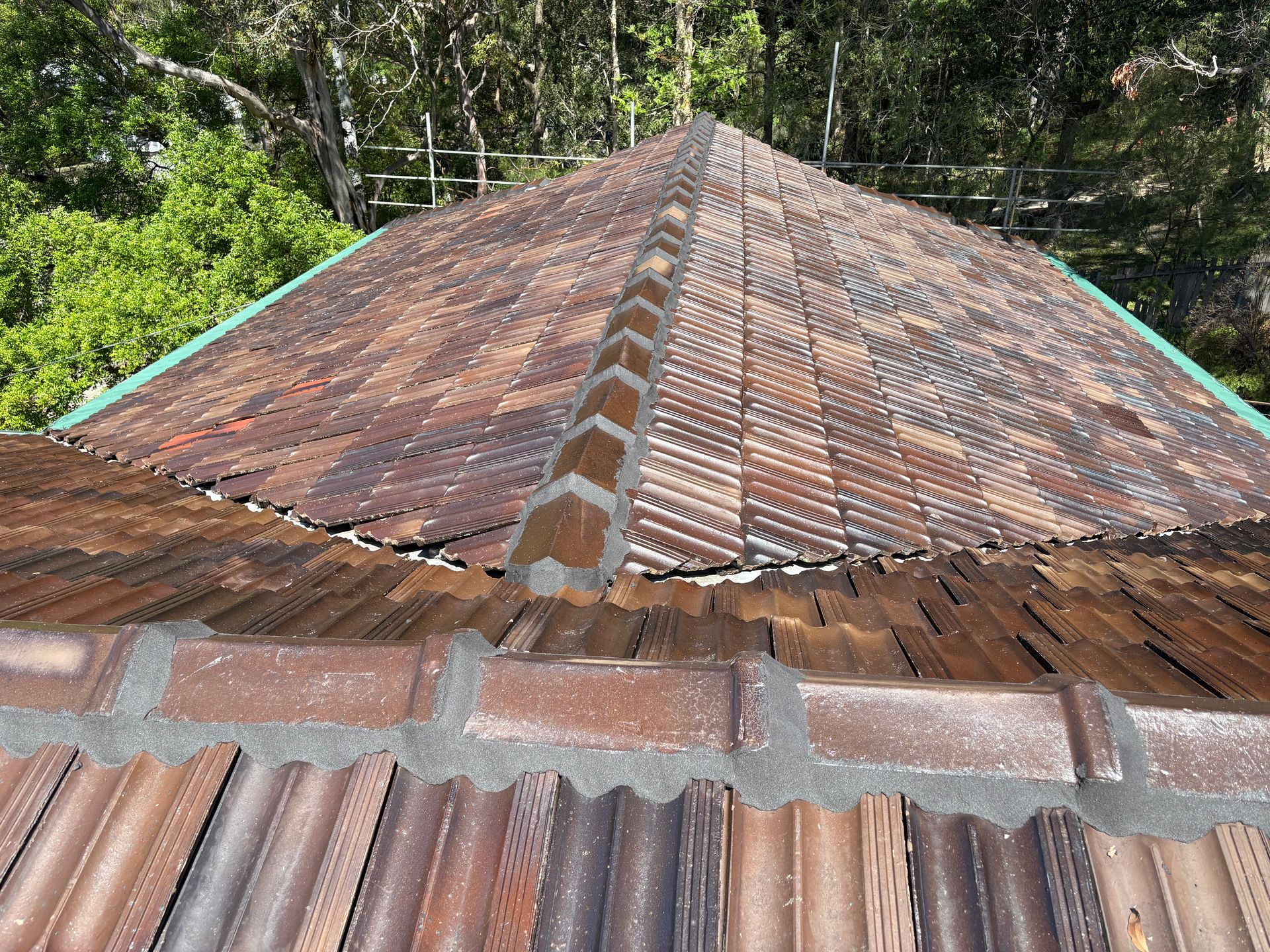 Rusty corrugated metal roof with a central ridge, viewed from above, surrounded by green trees — In-Time Roofing In Newcastle, NSW