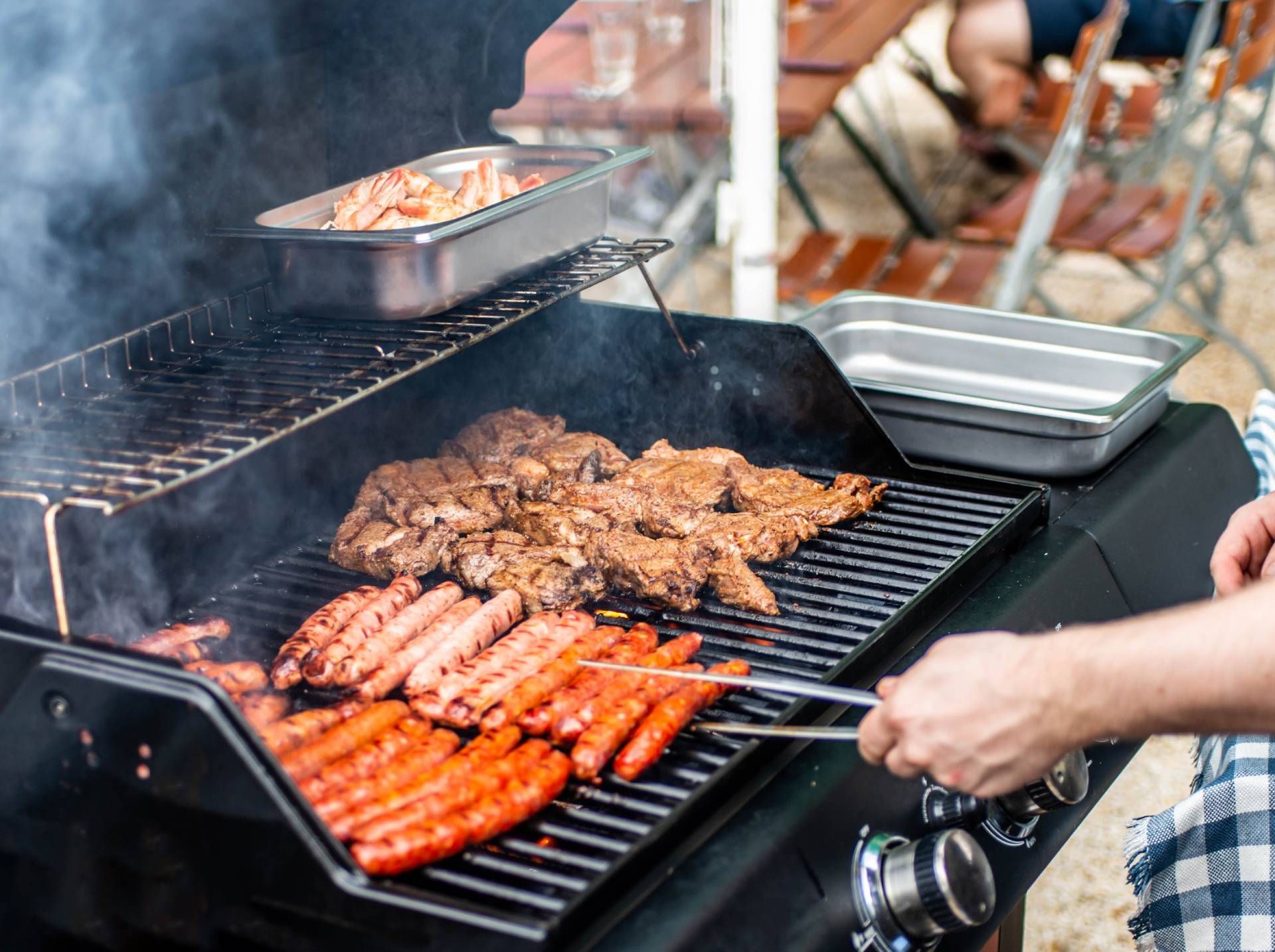 Person grilling skewers and meat on a smoky outdoor barbecue, with trays and a hand turning food on the grill