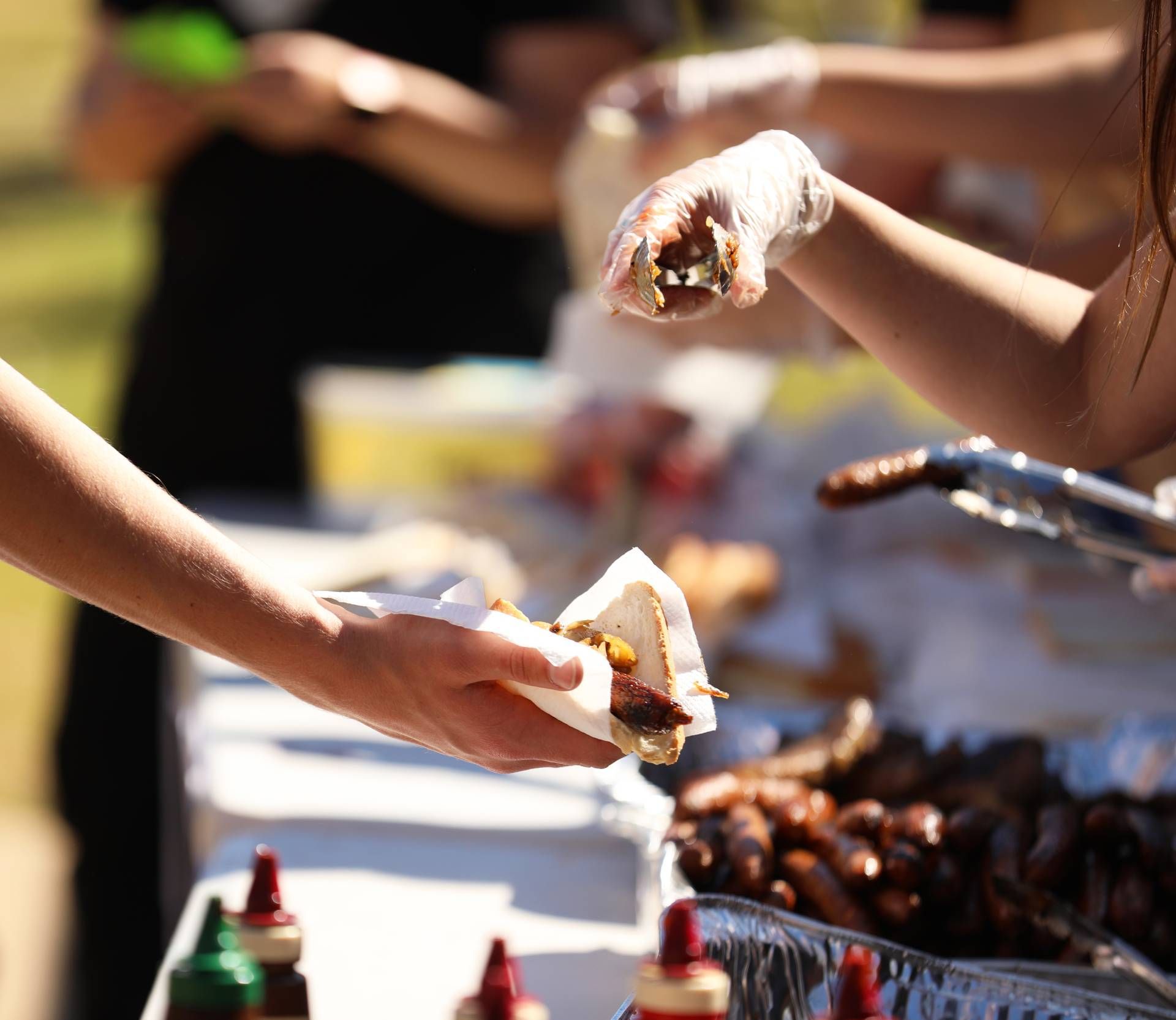 Hands serving barbecue food at an outdoor buffet with tongs and sauce bottles