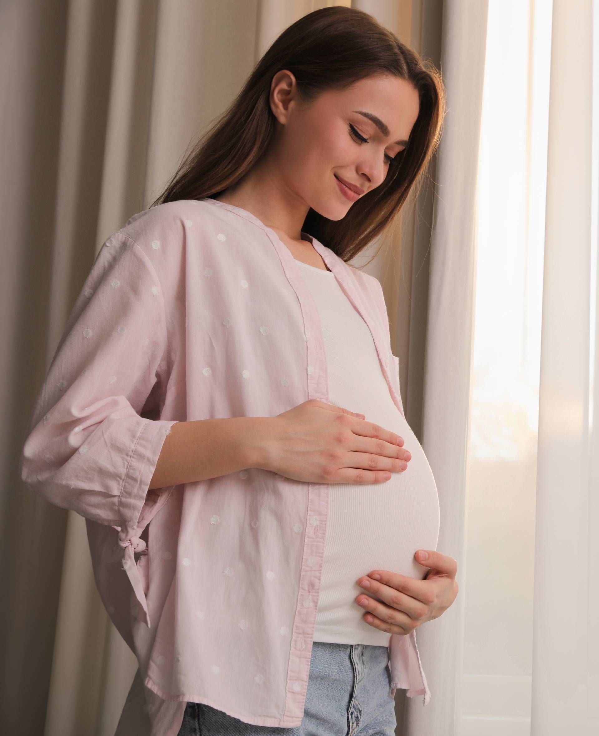 Pregnant woman in a pink blouse gently holding her belly by a sunlit window