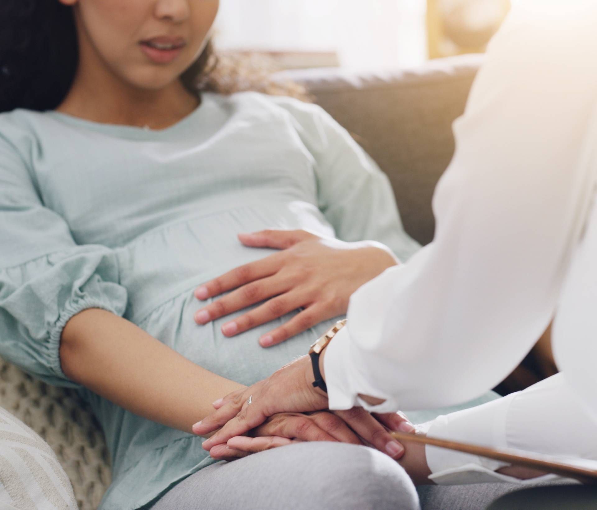 Pregnant person sitting on a couch, holding their belly while another person comforts them by hand