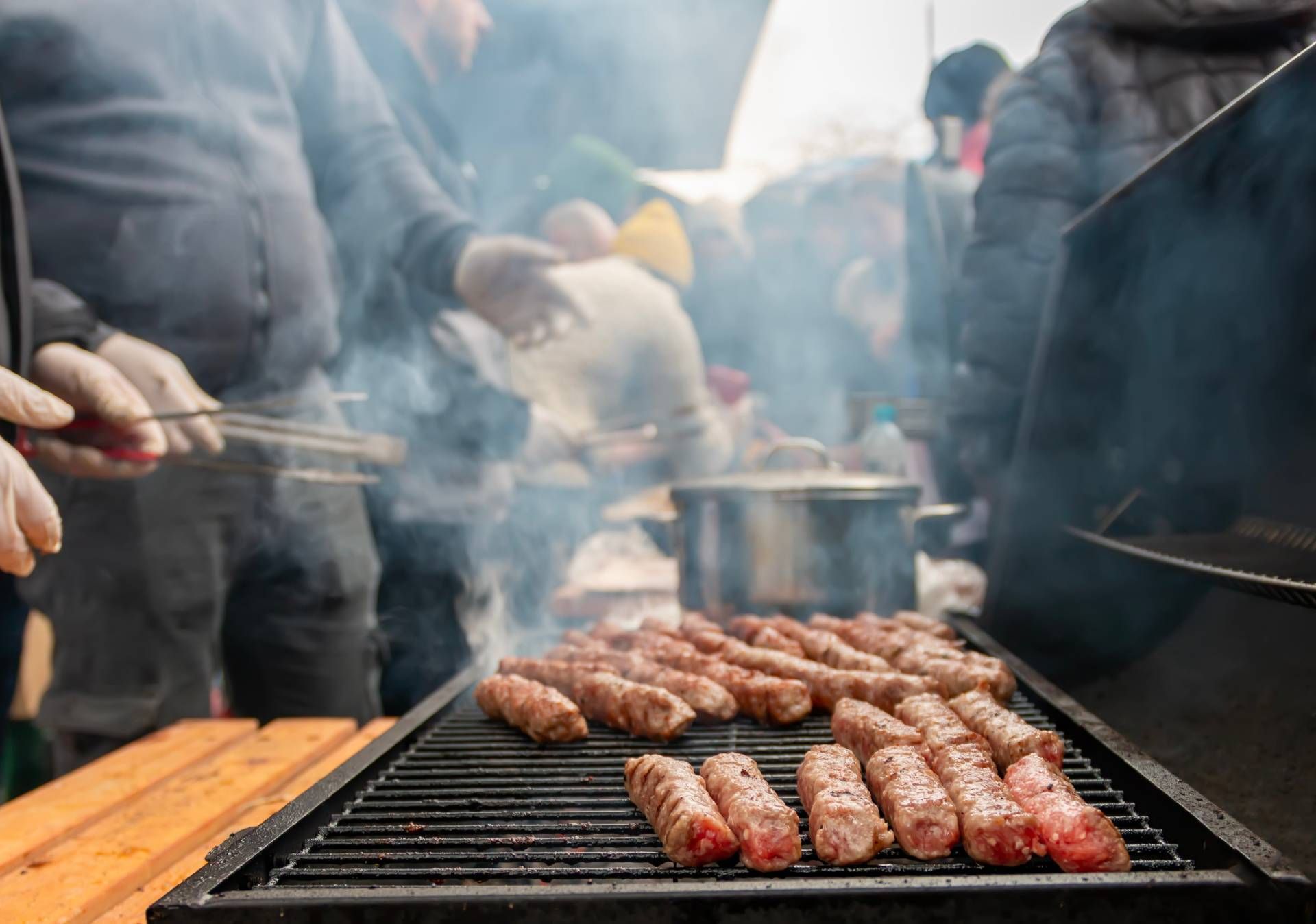 Grilled sausages sizzling on a barbecue grill amid smoke and gathered people