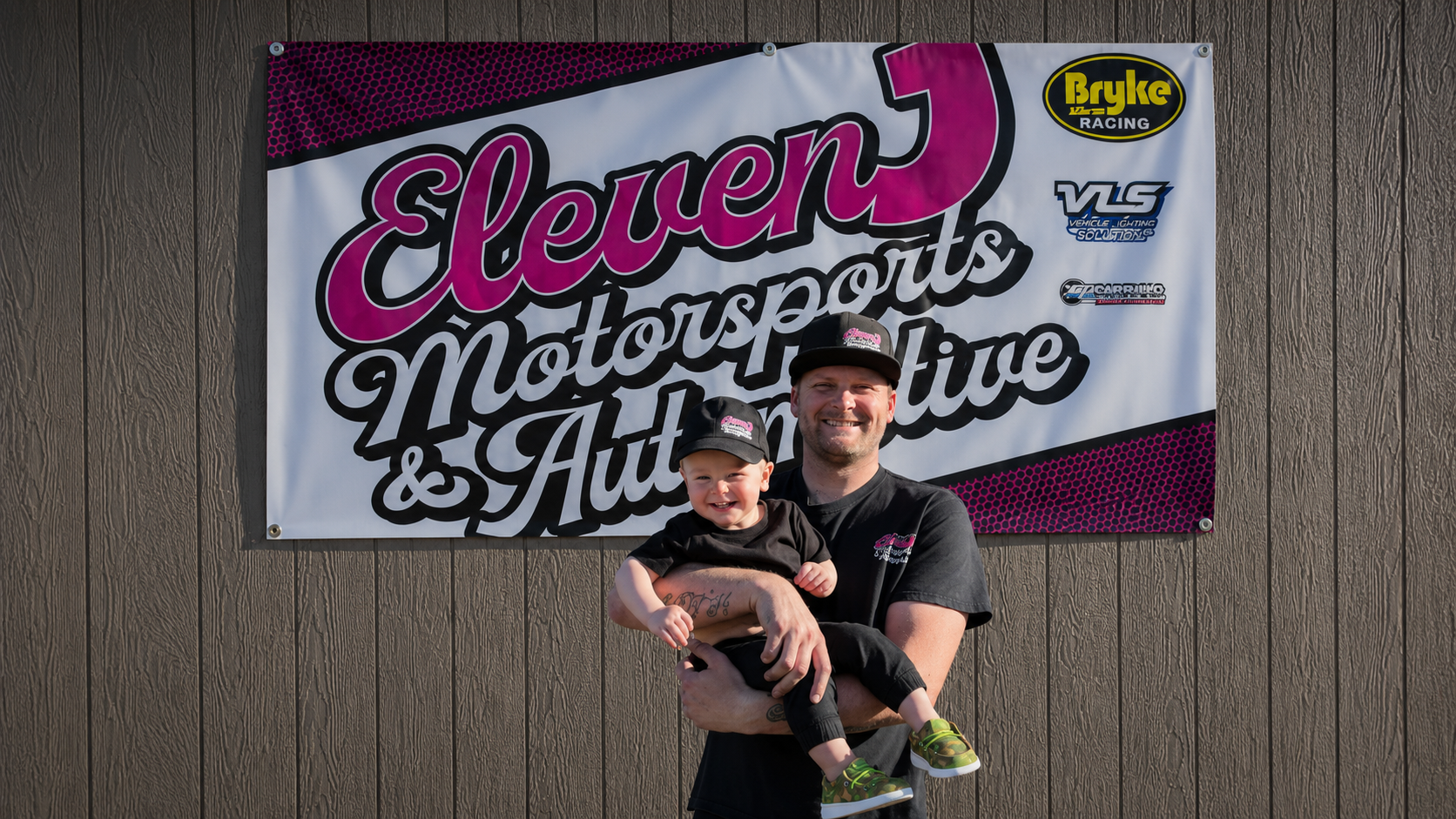 Two smiling people holding a baby in front of an Eleven Motorsports & Auto Service sign