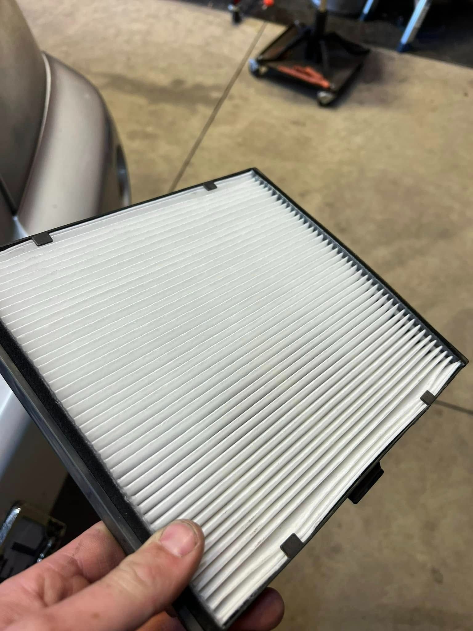 Hand holding a white pleated cabin air filter with a black frame in a garage.