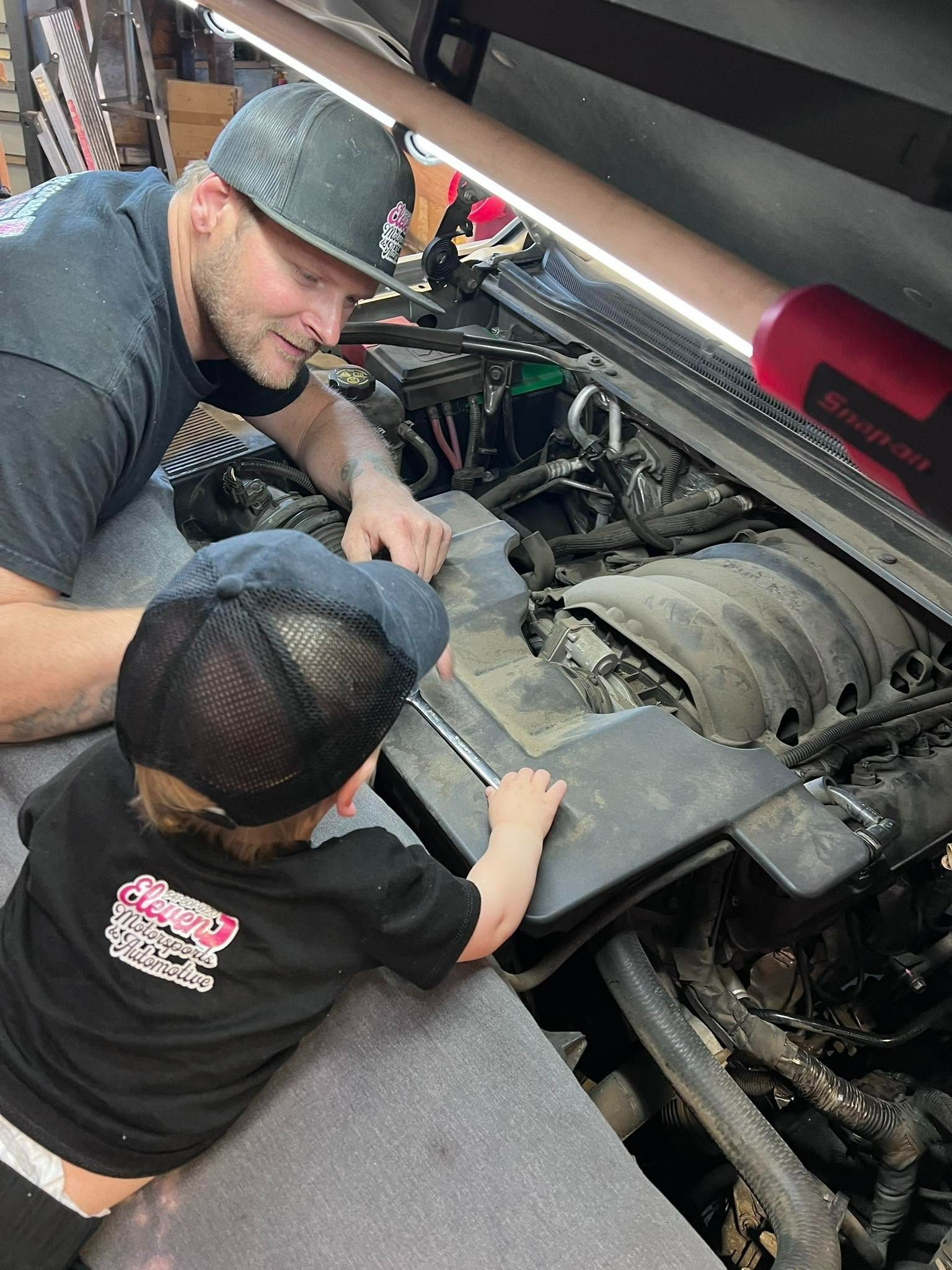 Two people inspect an engine under a raised hood in a garage.