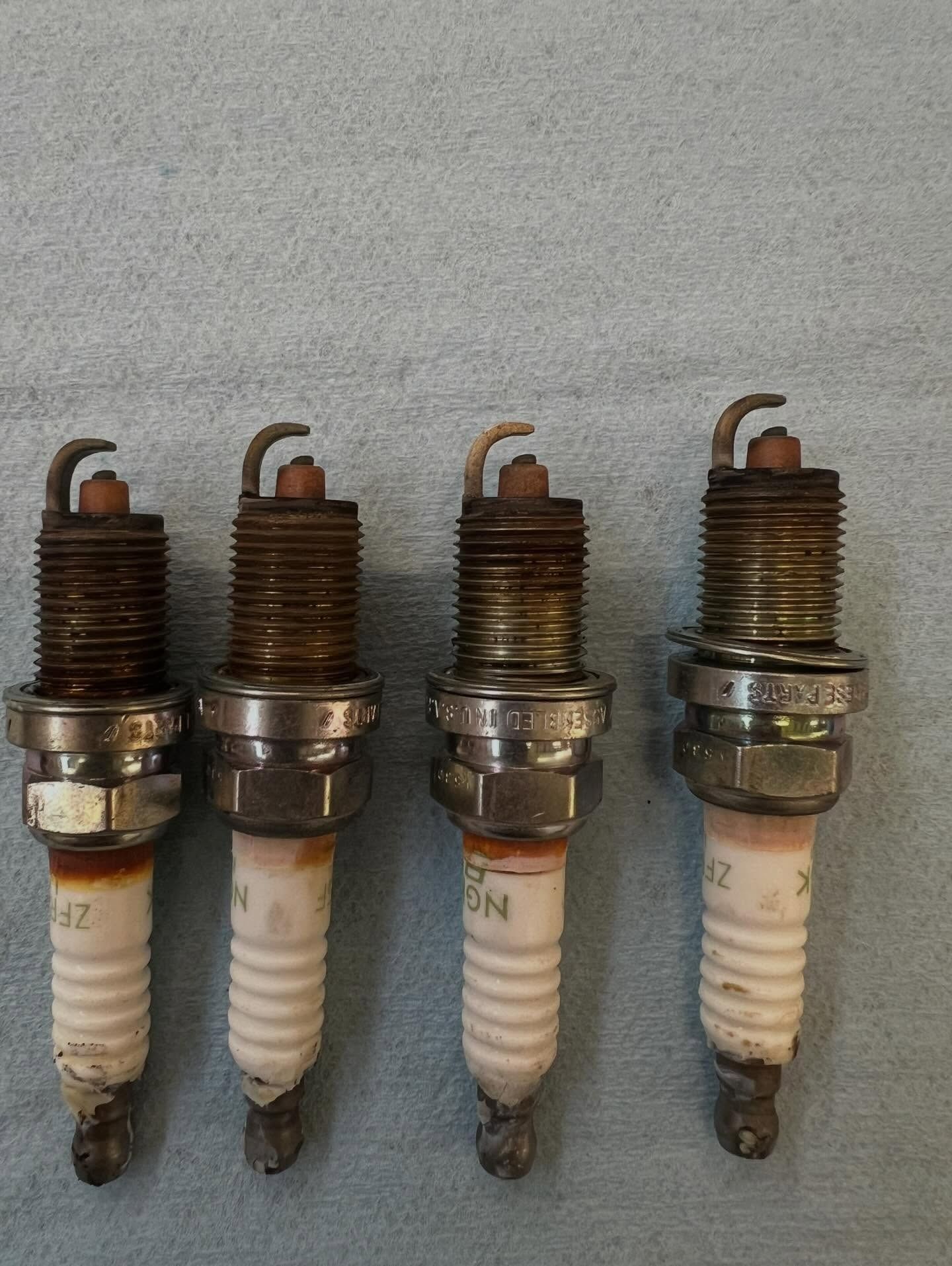 Four used spark plugs lined up on a gray surface, showing dark wear on the tips.