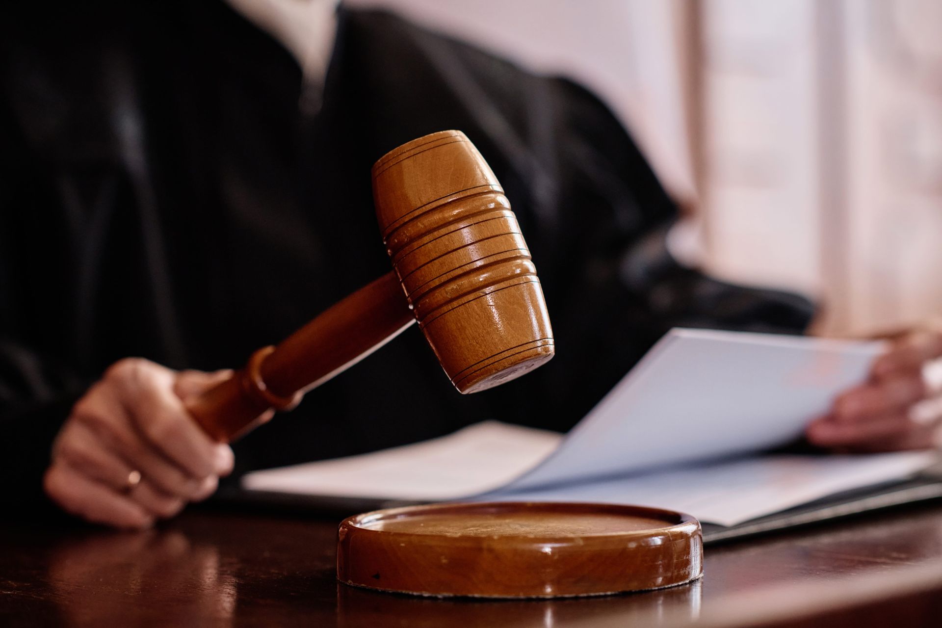 Judge’s gavel above a wooden sound block in a courtroom, with a person reading documents behind it.