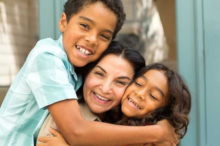 Woman and two children embrace, smiling outdoors.