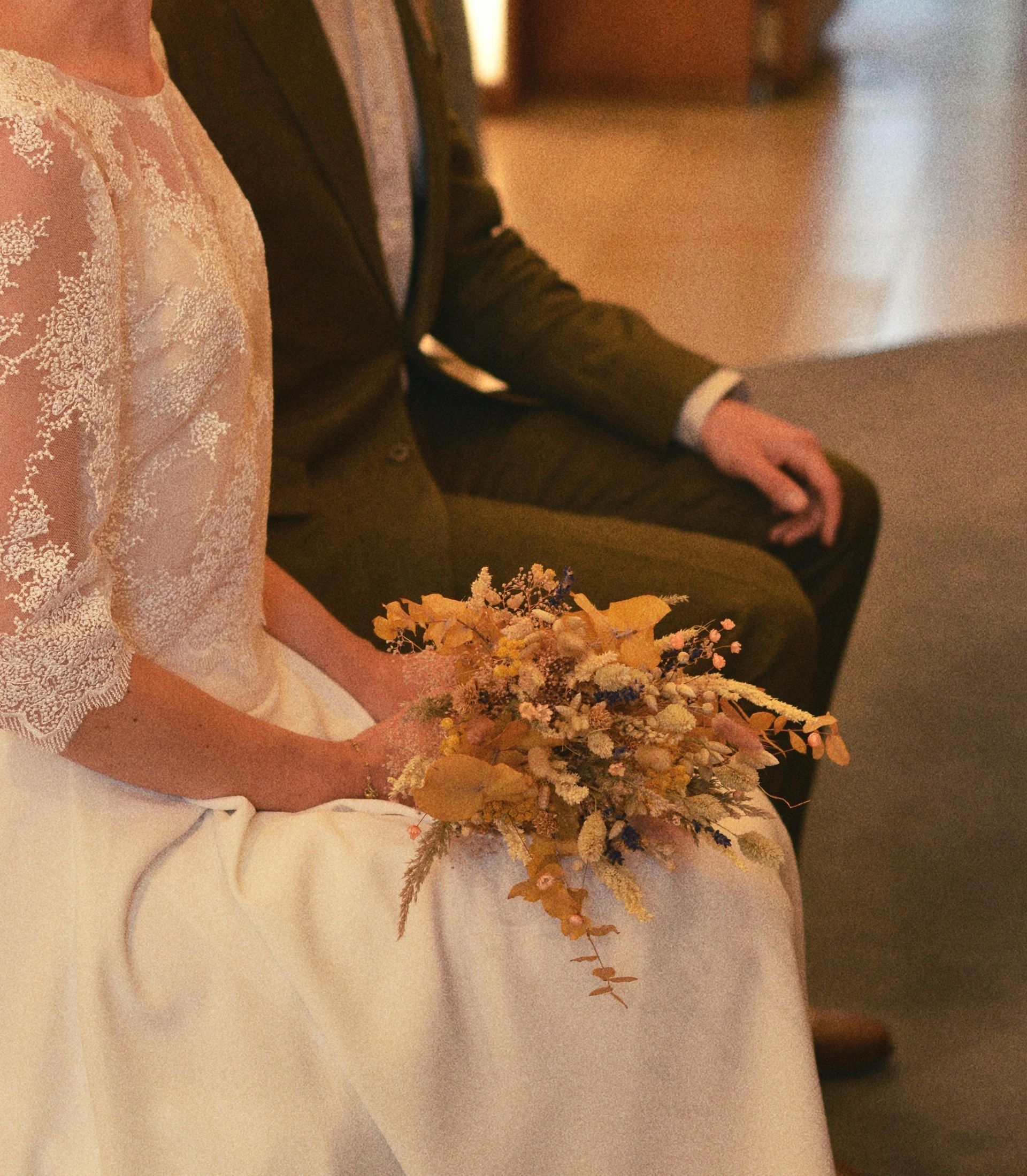 A person in a lace wedding dress holding a dried-flower bouquet sits next to someone in a green suit.