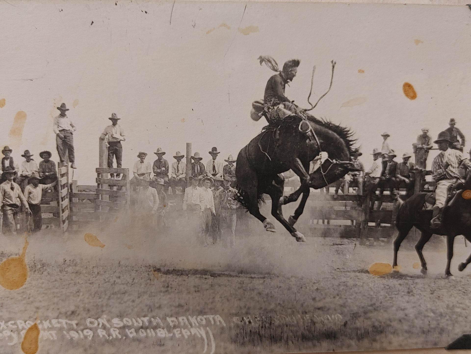 Bronco rider mid-jump in dusty rodeo arena, with spectators and fence in the background