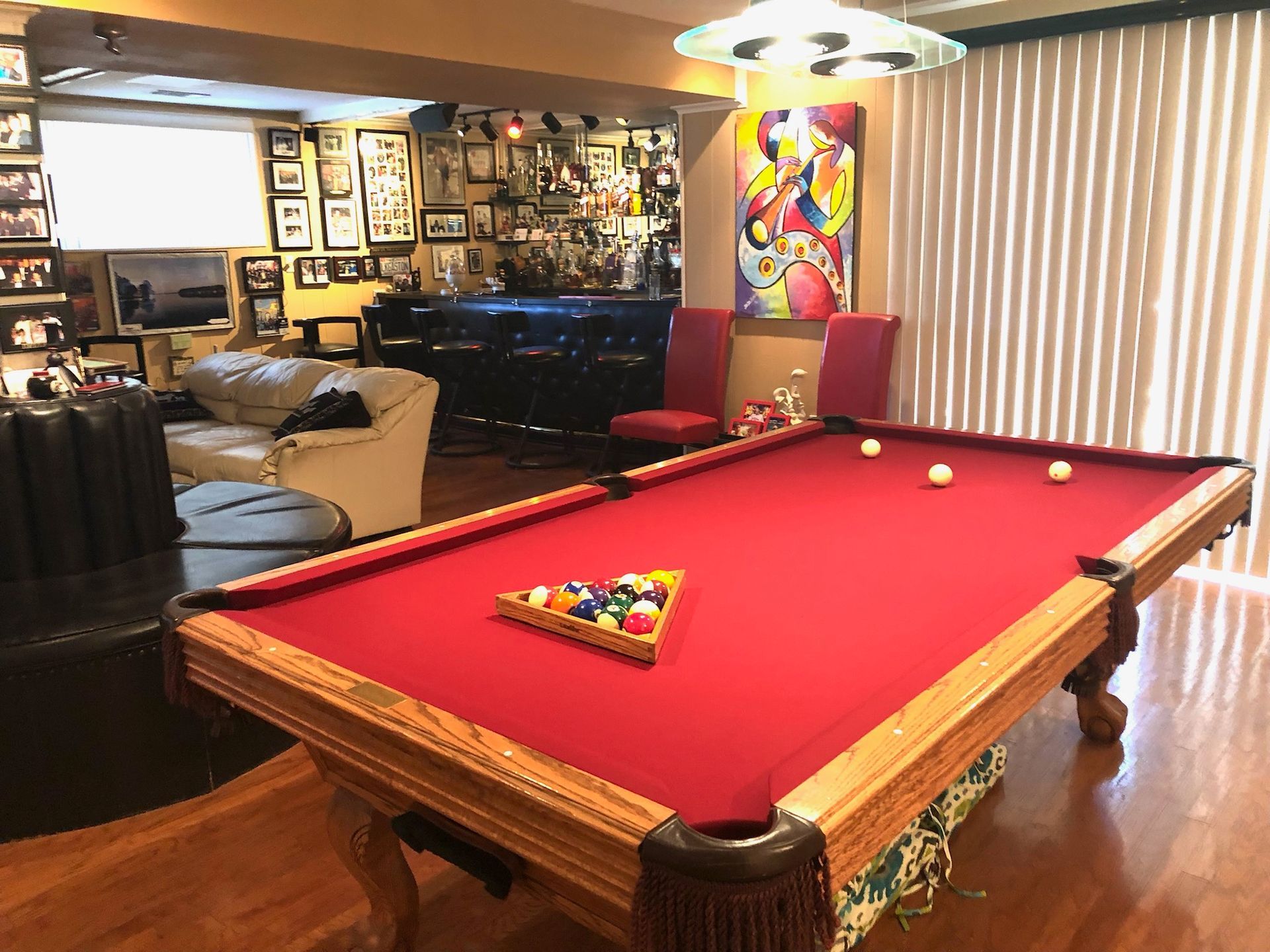 Red-felt pool table in a cozy game room with balls racked and scattered, bar area in the background