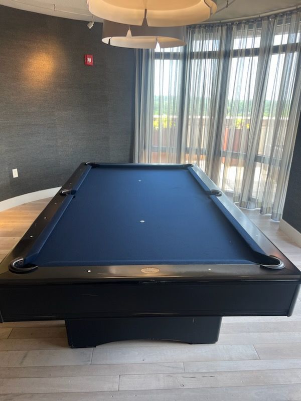 Blue pool table in a modern room with dark walls and large windows with sheer curtains.