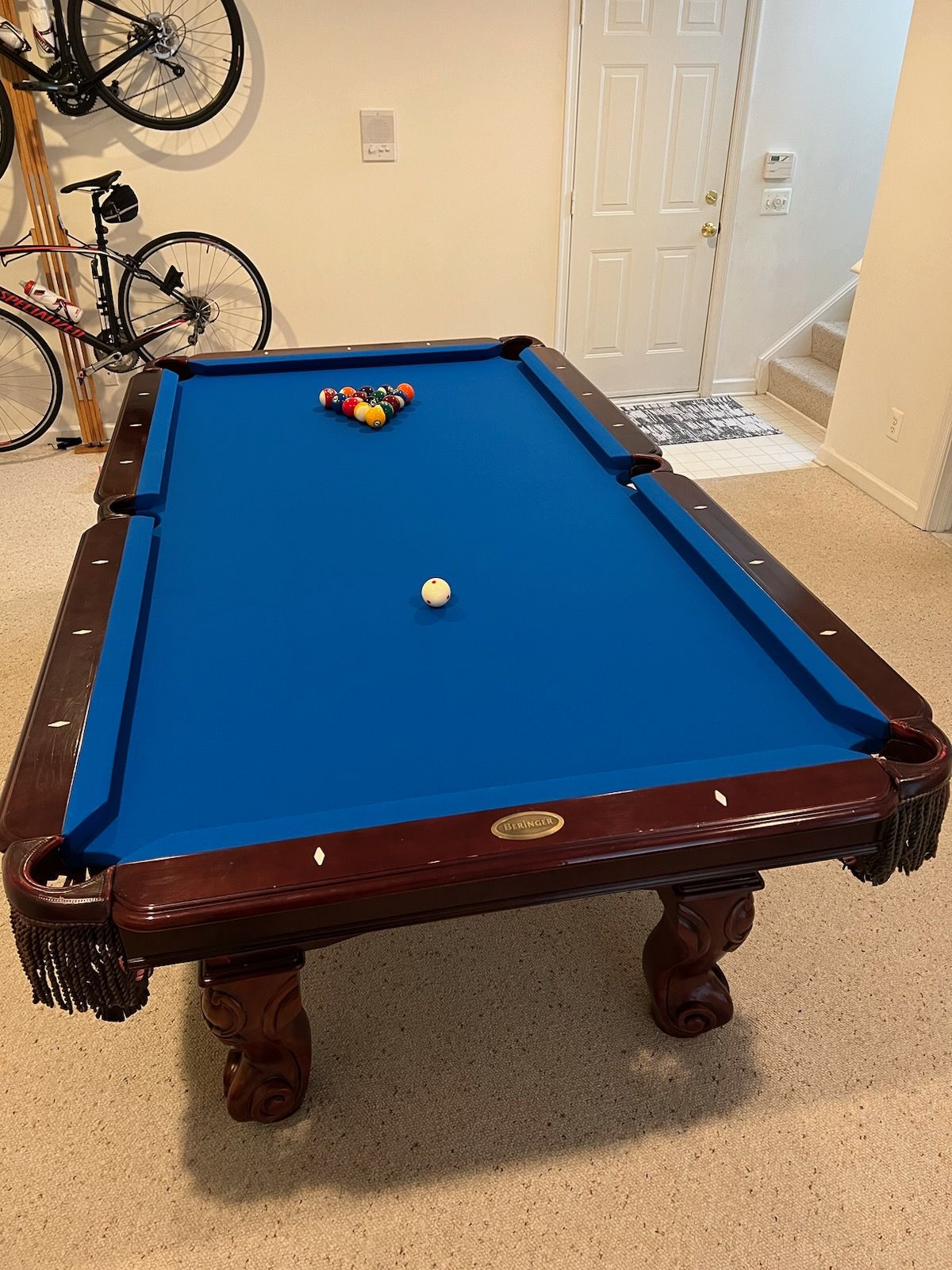 Blue pool table with one white ball and a rack of balls in a room with bicycles and a door.