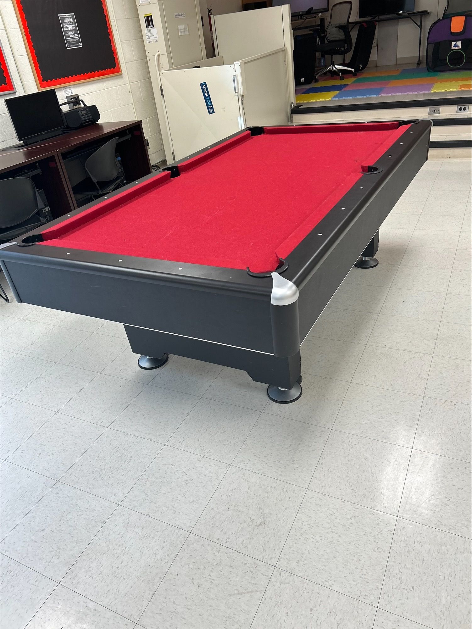 Red-felt pool table in a game room with cabinets and shelves in the background.