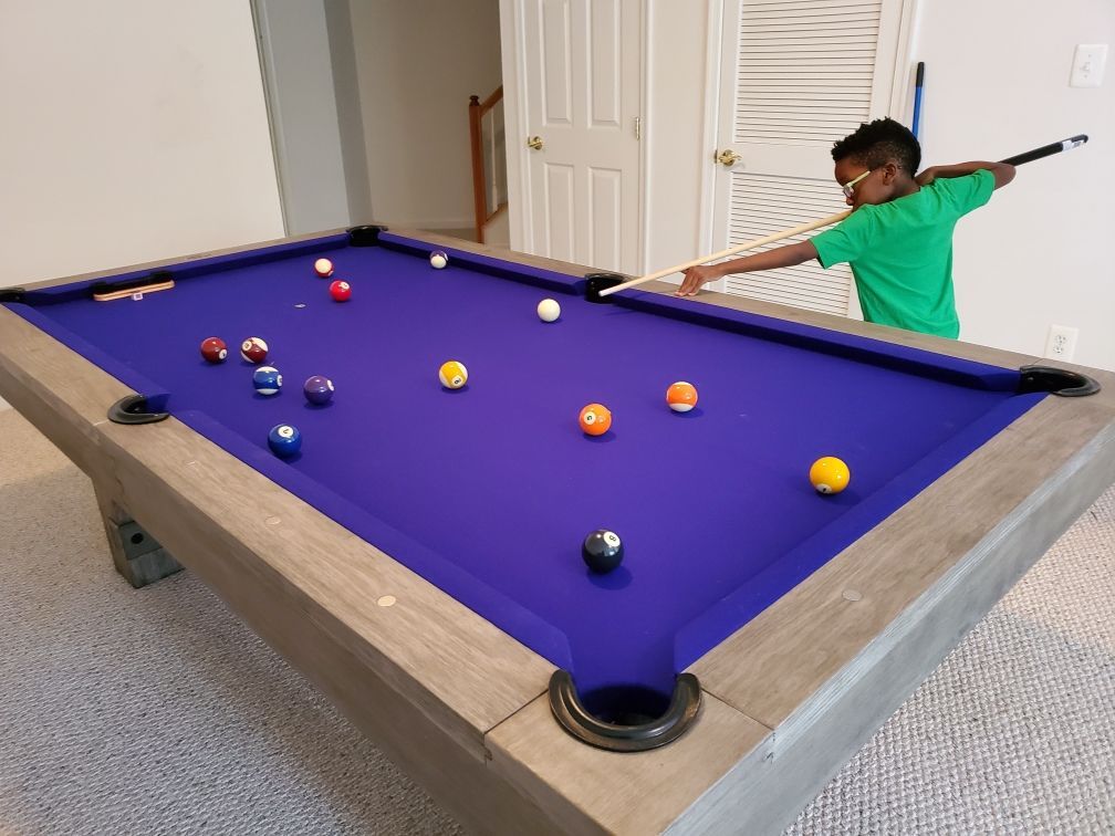 Child aiming a cue stick at a purple pool table with scattered billiard balls in a carpeted room