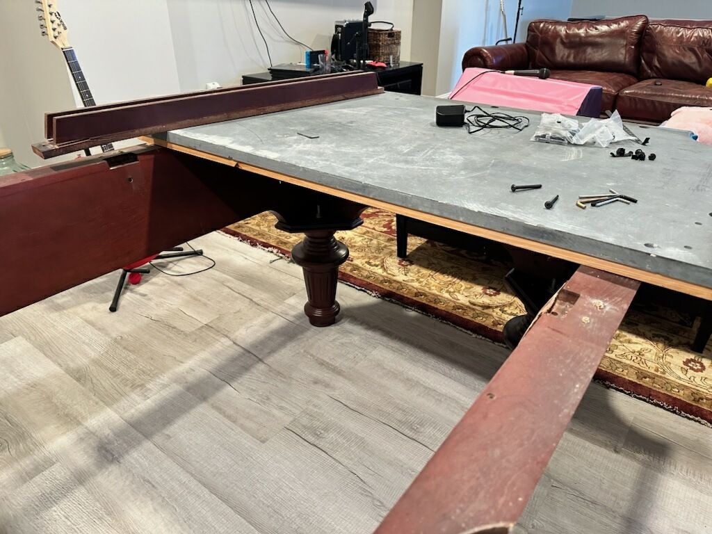 Pool table under repair with removed rails and tools on a gray felt surface in a living room