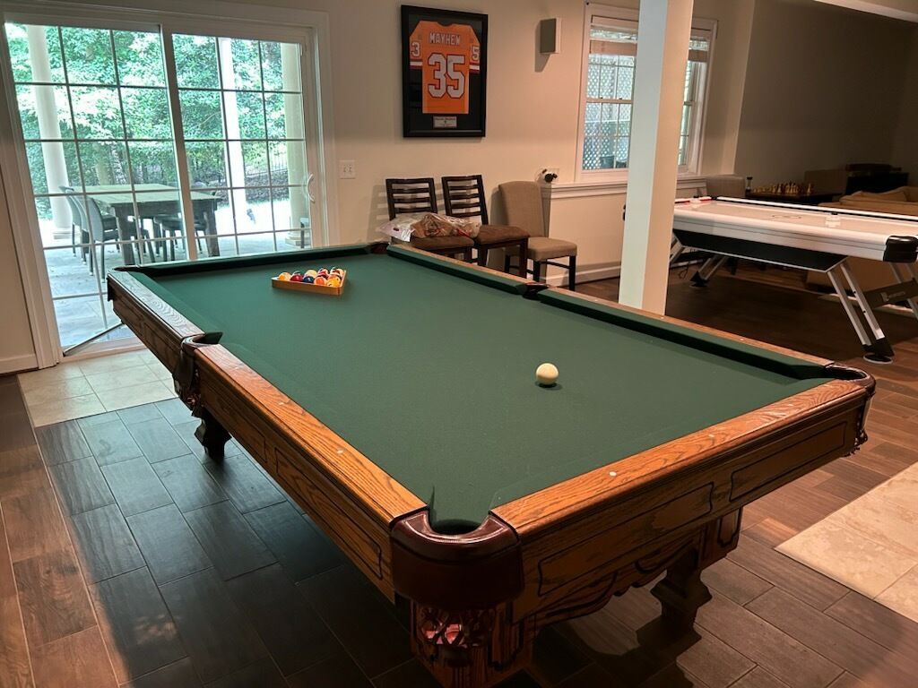 Pool table in a bright room with large windows, a framed jersey on the wall, and lounge seating nearby.