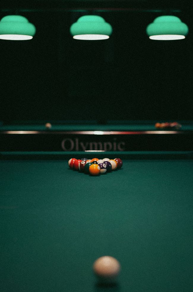 Pool table with racked balls at the far end and a cue ball in the foreground under green hanging lights