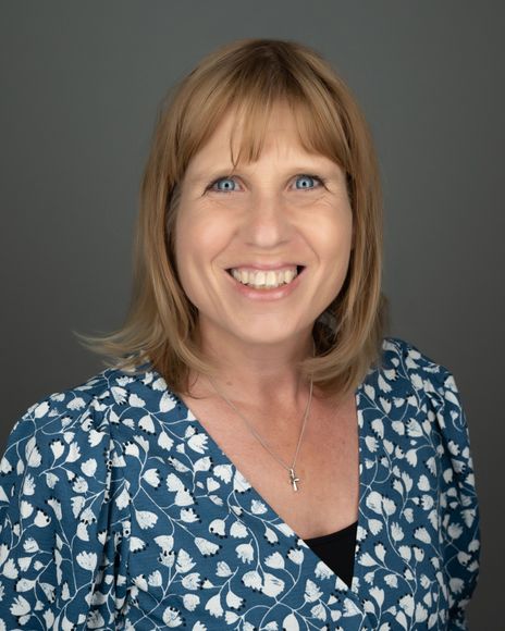 Woman with blonde hair and blue eyes, smiling, wearing a blue floral patterned top, against a gray backdrop.