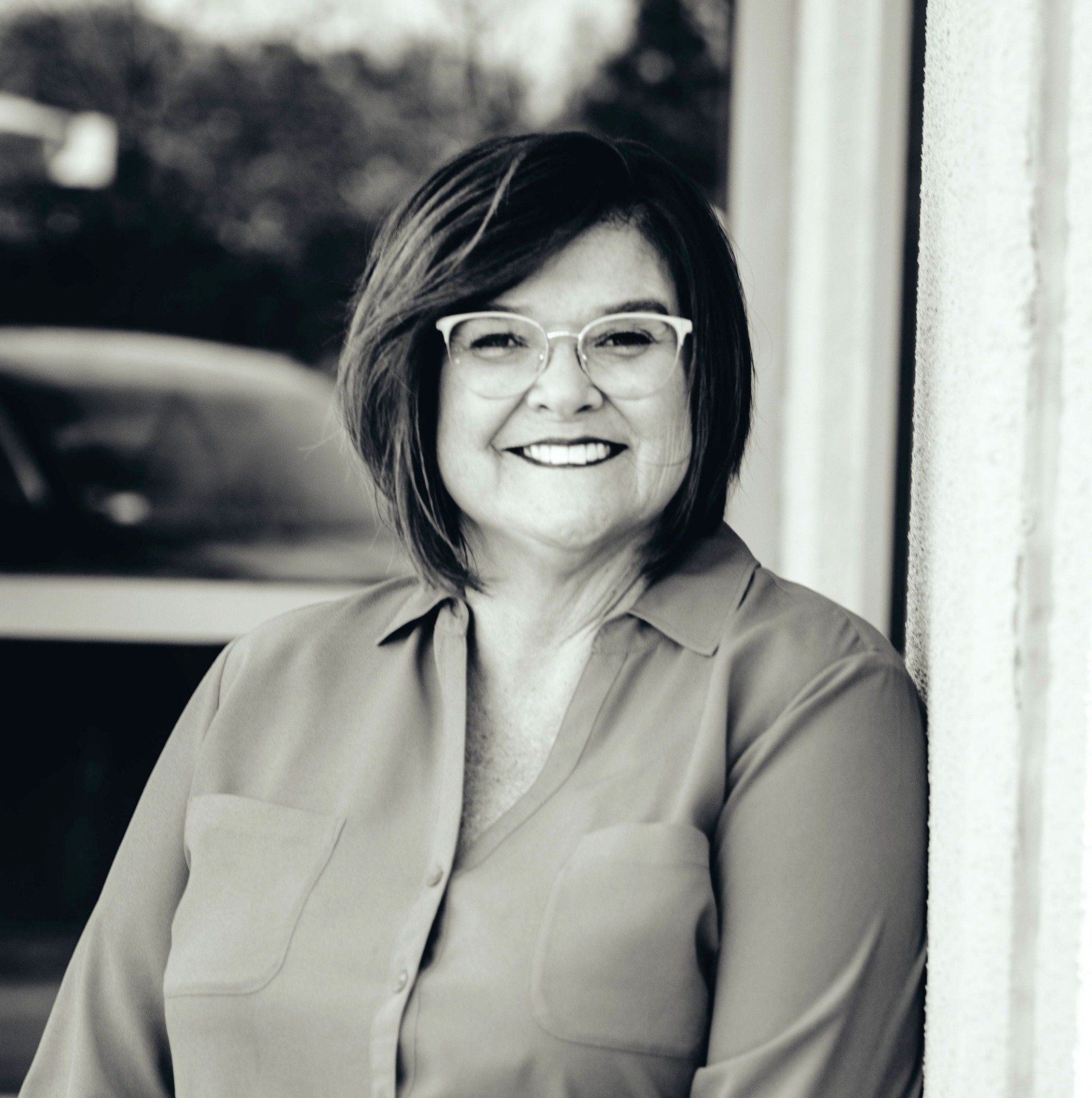 Woman with glasses smiles while leaning against a white textured wall outside.
