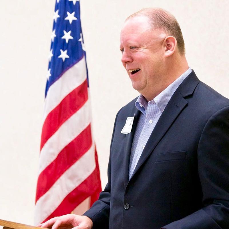 Man in suit smiles at a podium, American flag in background.