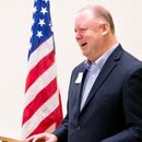 Man in a suit smiles, standing near the American flag.