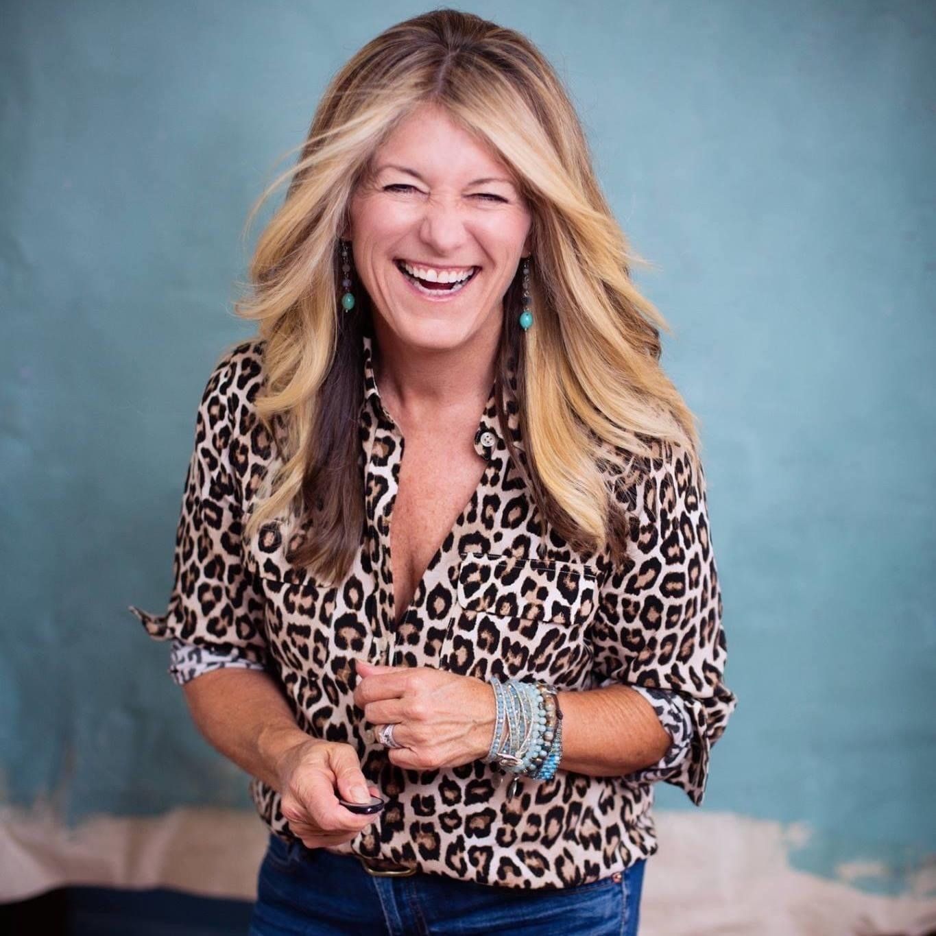 Woman laughing, wearing leopard print shirt and turquoise earrings, in front of a blue wall.